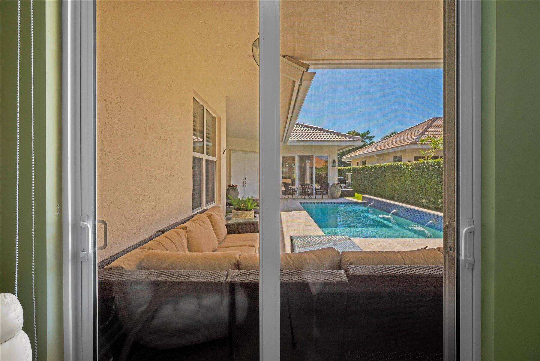 Backyard pool and covered patio viewed through sliding glass doors, with wicker seating in the foreground.