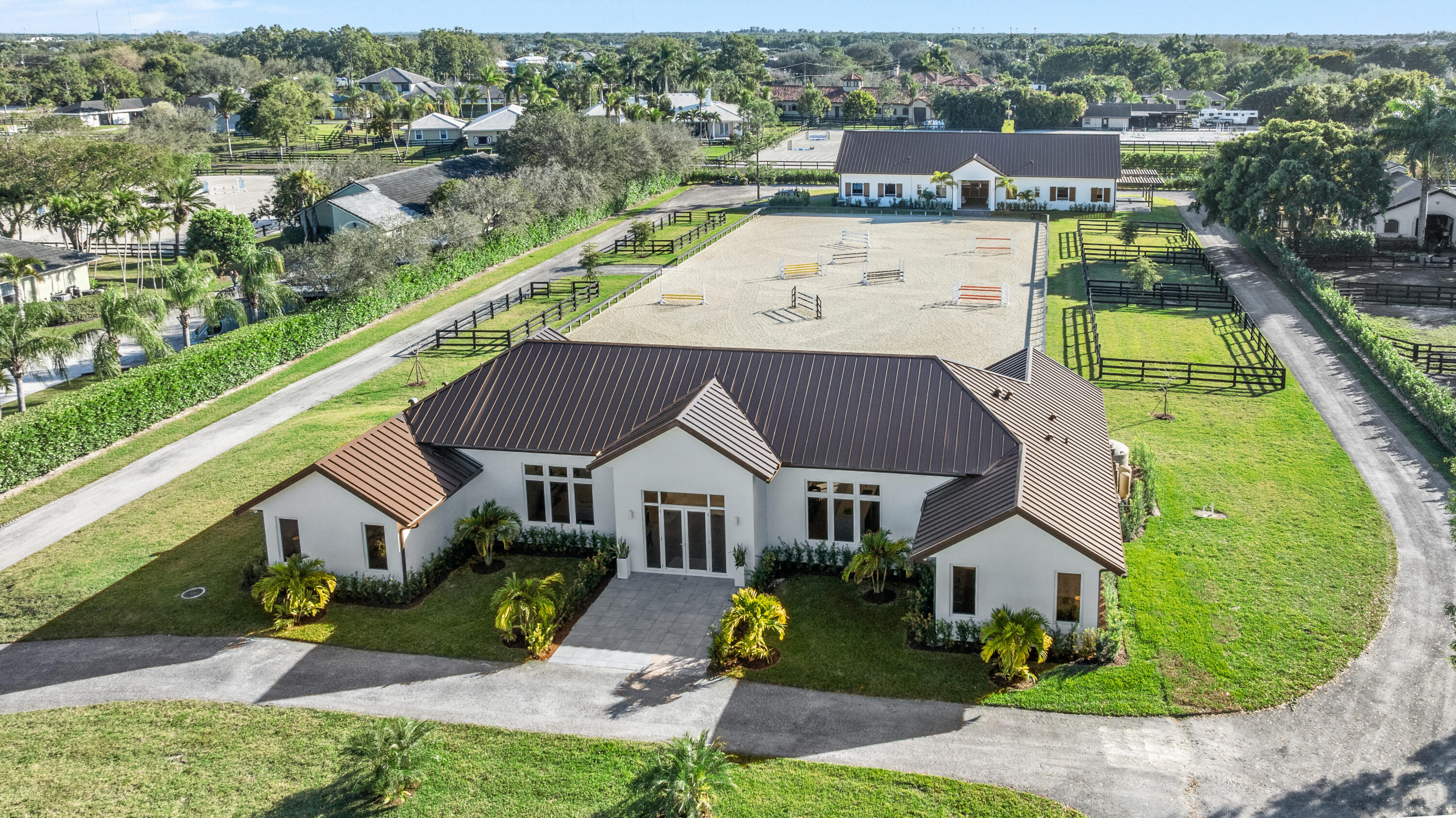 Aerial view of a white home beside a fenced riding arena with jump obstacles and paddocks.
