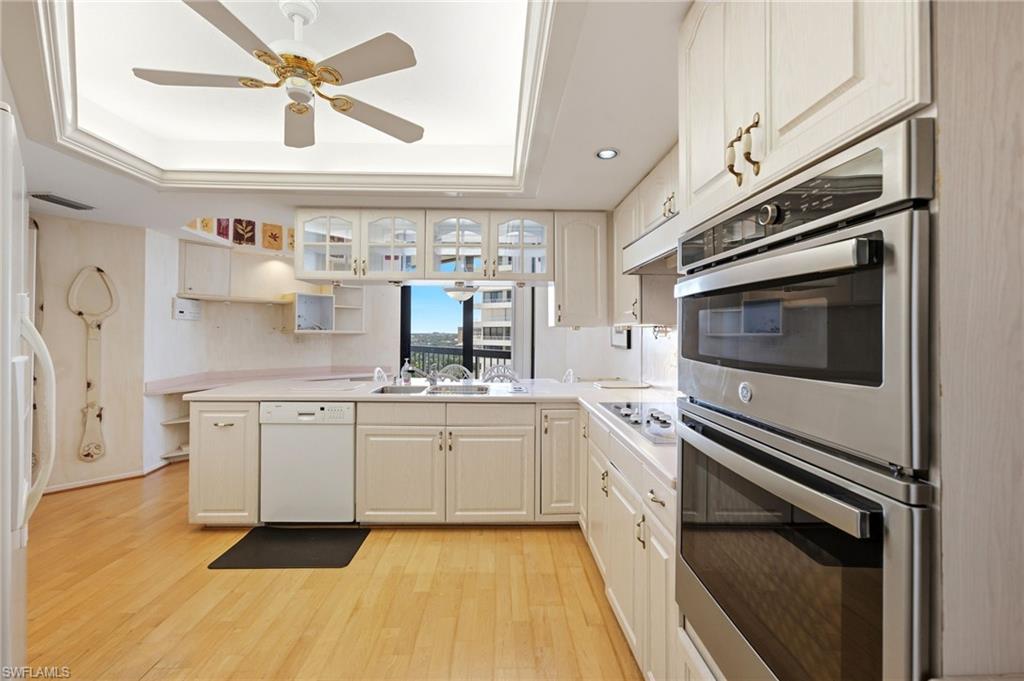 White kitchen with stainless steel double ovens, glass-front cabinets, and a sink beneath the window.