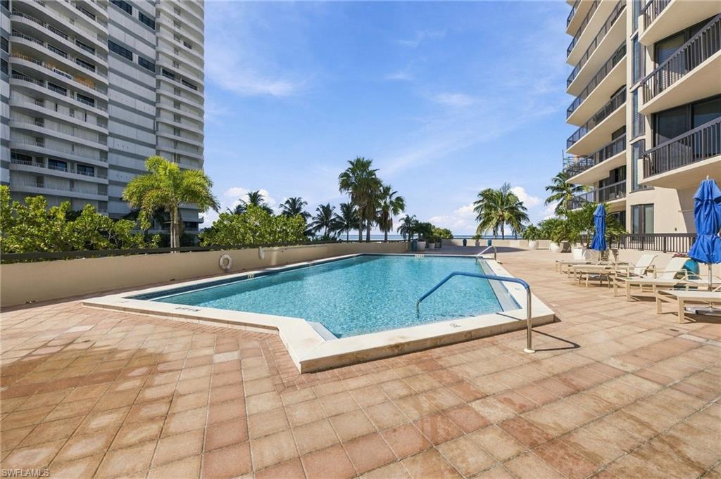 Condominium pool between high-rises, with palm trees and a glimpse of water beyond.