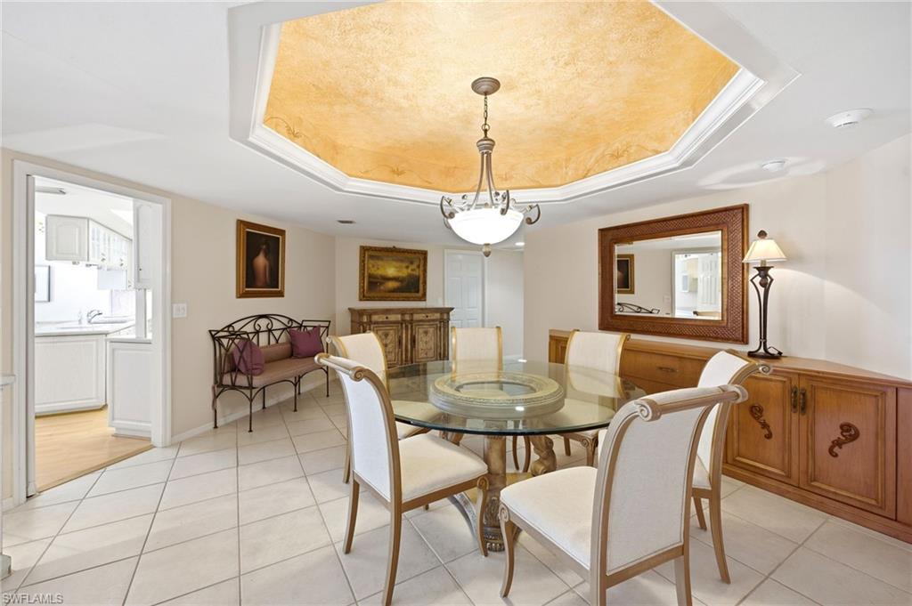 Dining room with a glass-top table, chandelier, gold-toned tray ceiling, and wooden sideboard.