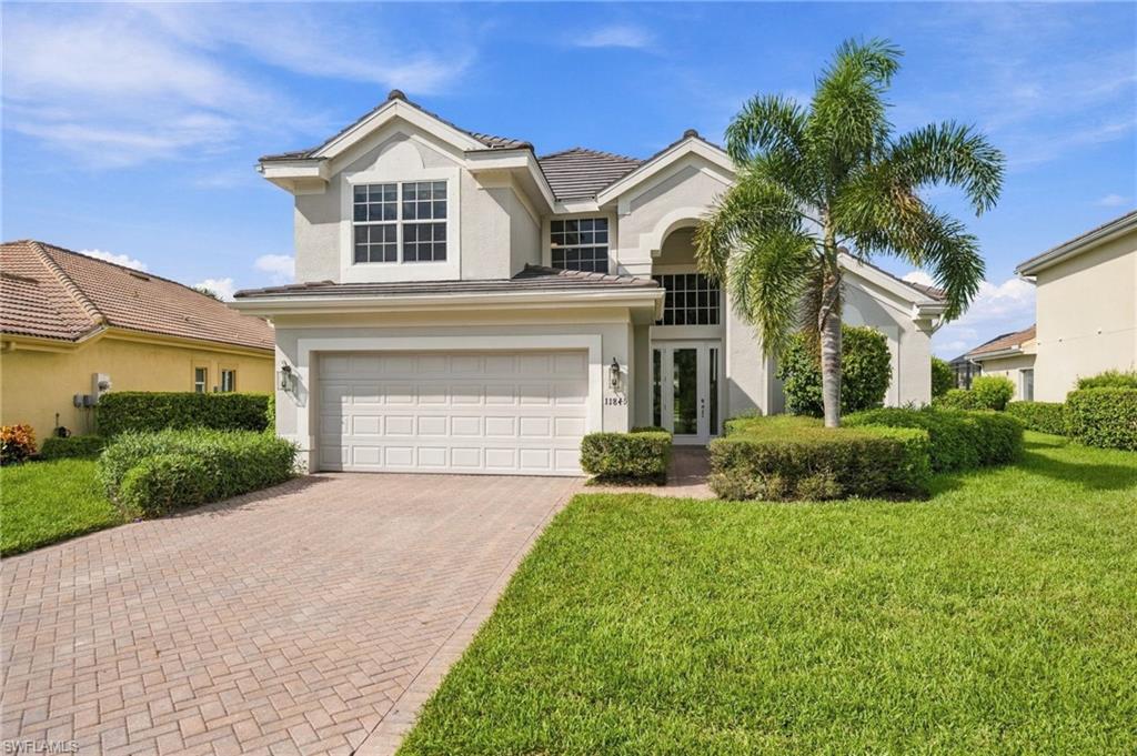 Two-story stucco home with a double garage, paver driveway, palm trees, and a green lawn.