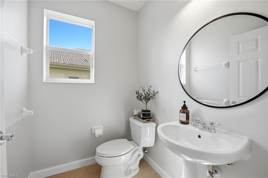 Bathroom with a pedestal sink, round black-framed mirror, toilet, and window.