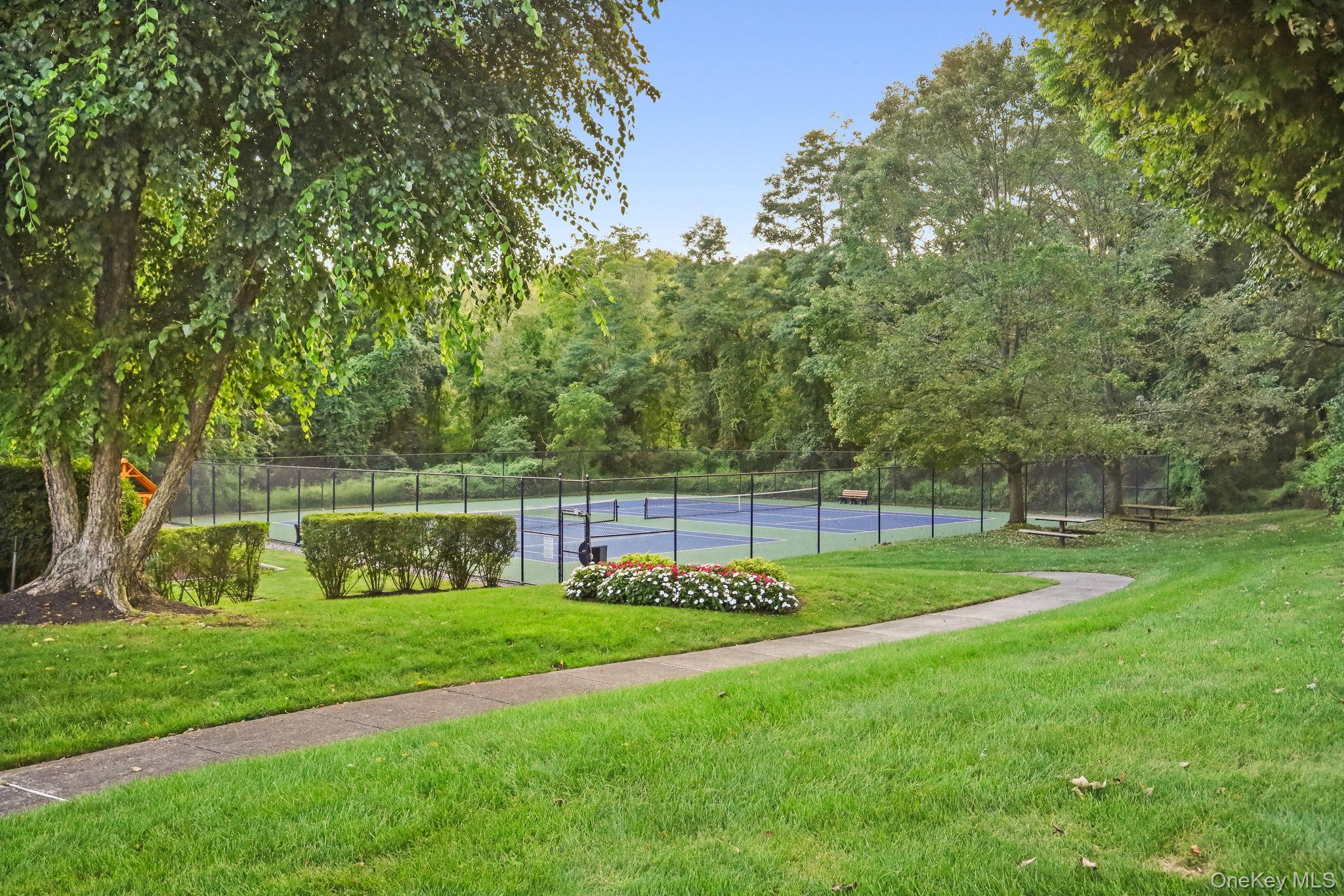 Fenced tennis court among mature trees, with a flower bed and curving lawn walkway.