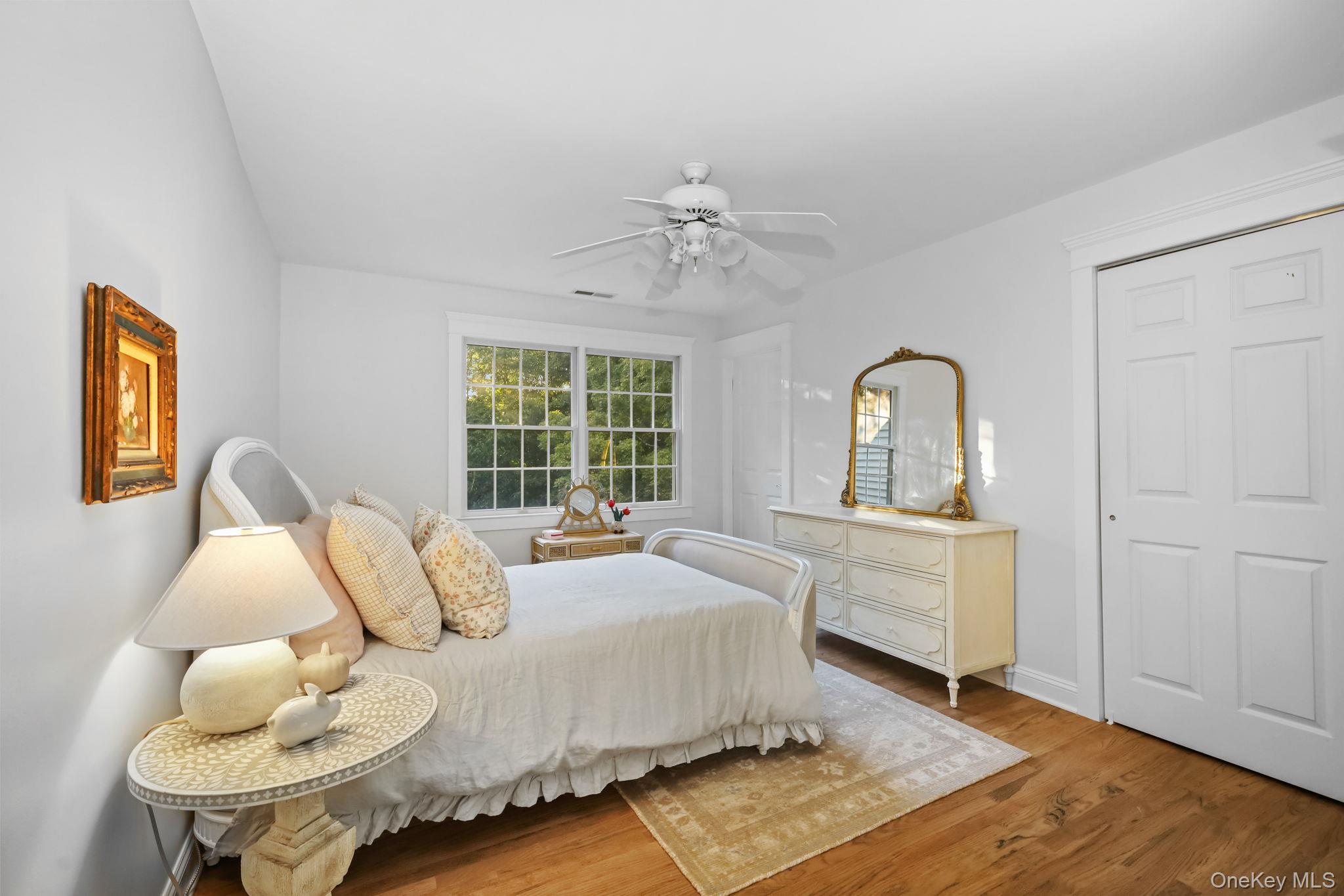 Furnished bedroom with a white bed, large double window, and cream dresser with a gold-framed mirror.