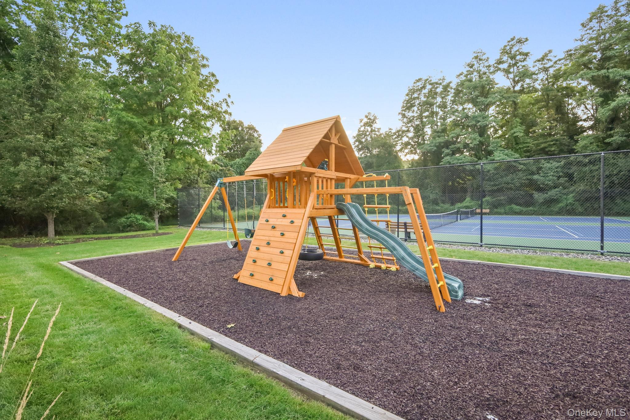 Wooden playground with swings, climbing wall, and slide beside a fenced tennis court