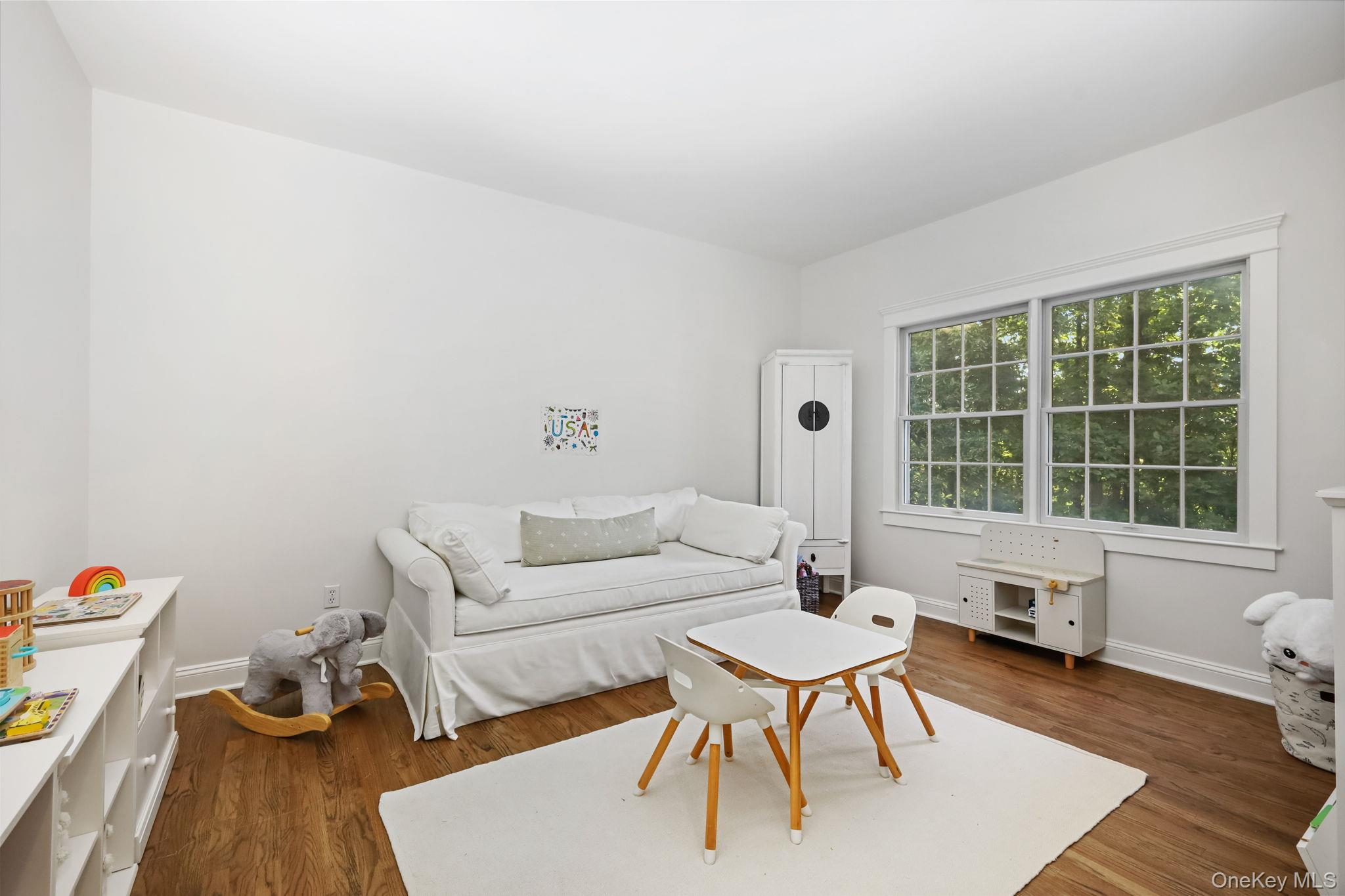Bright playroom with a white sofa, child-sized table and chairs, and a large window overlooking trees.