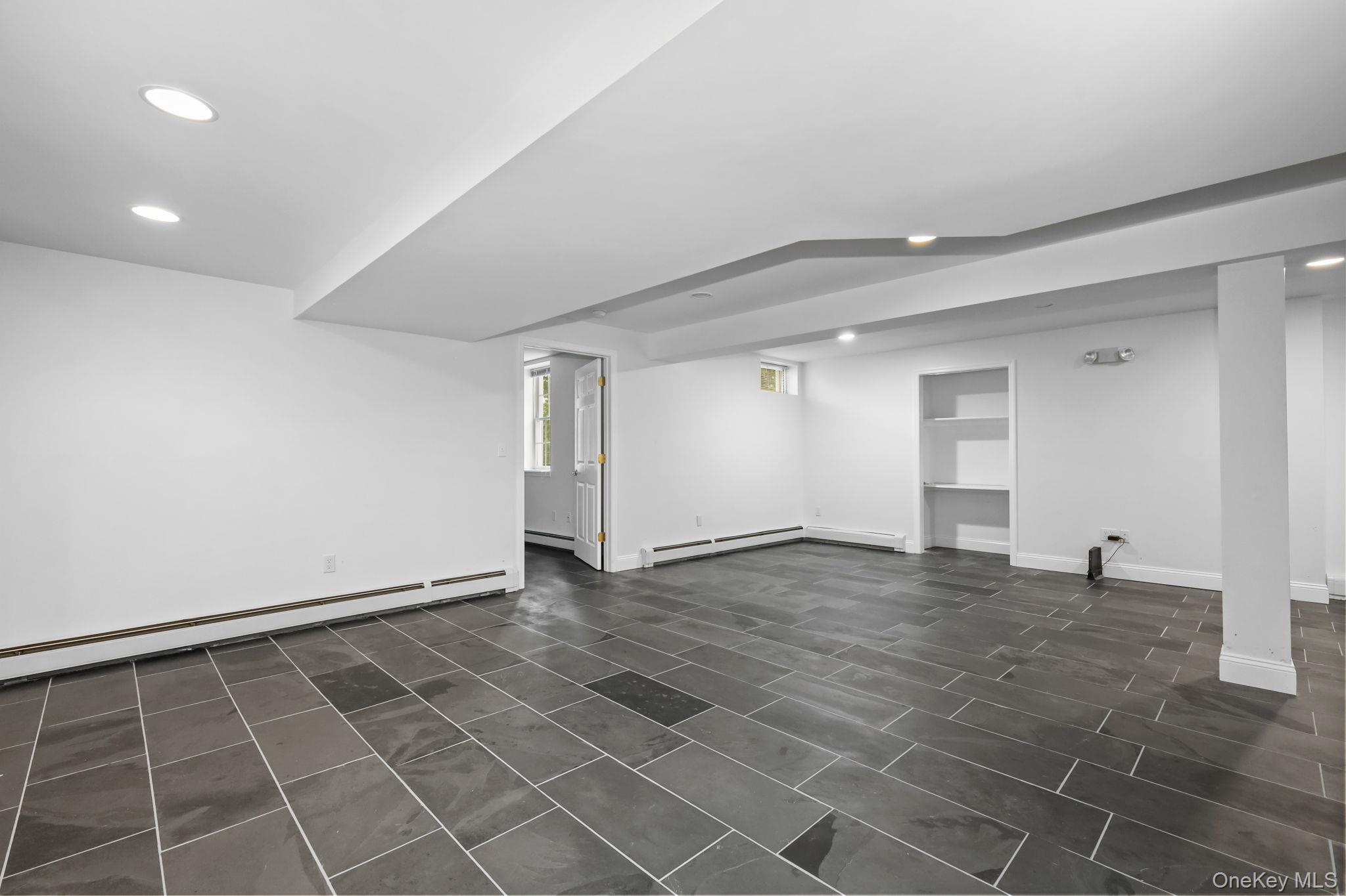 Empty basement room with large gray tile floors, recessed lights, and built-in shelving.