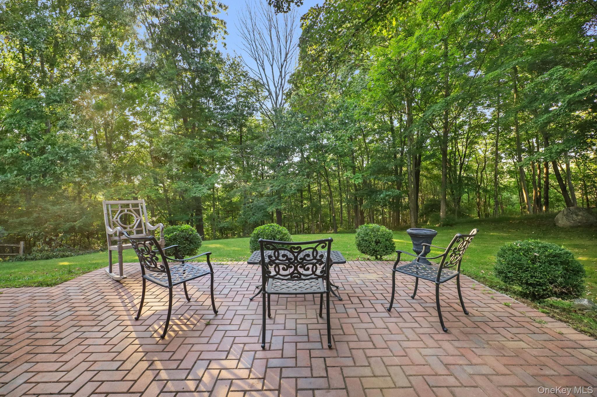 Brick patio with wrought-iron seating overlooks a landscaped lawn and wooded yard.