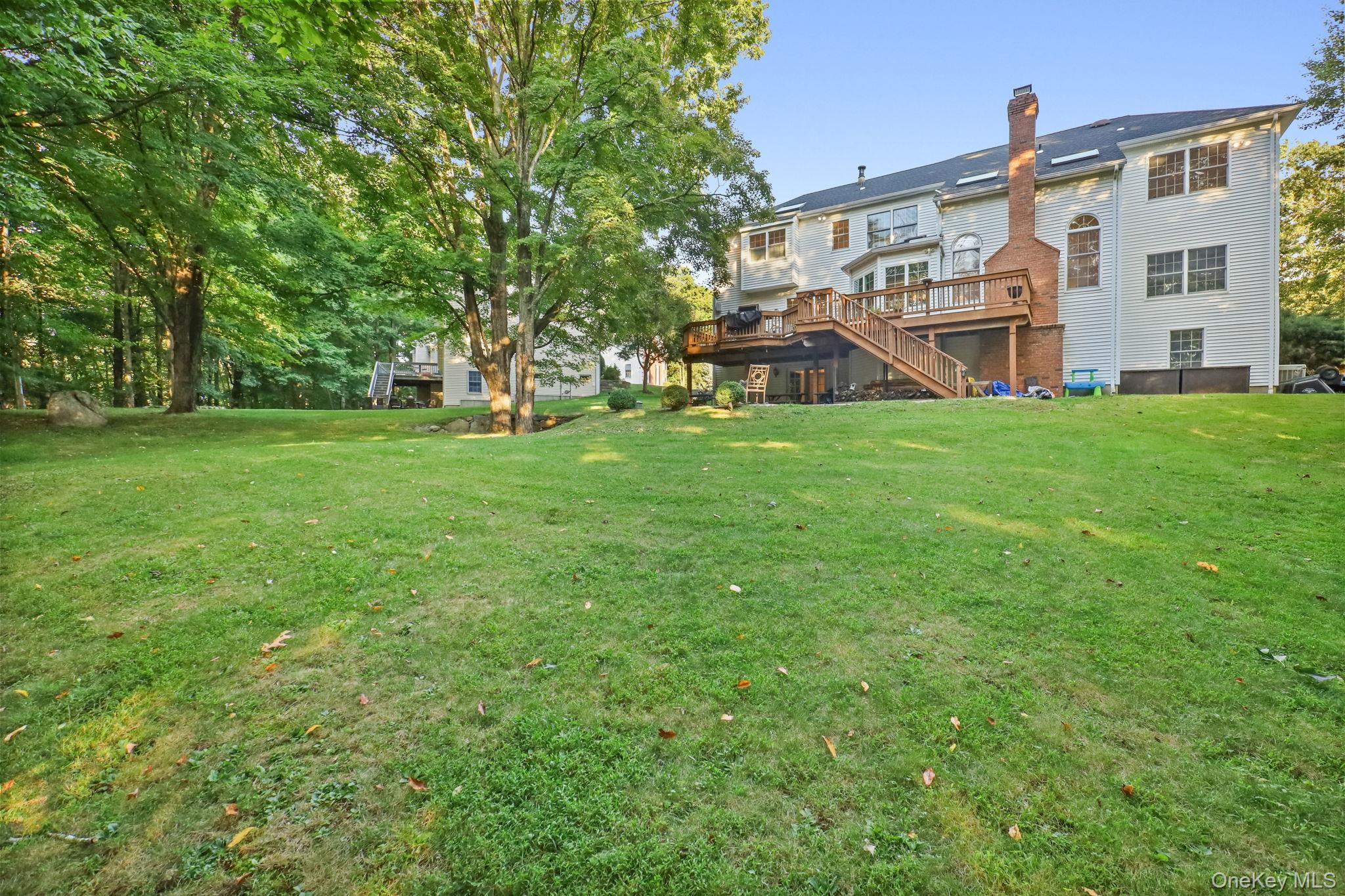 Rear of a white-sided house with a raised wooden deck, brick chimney, and broad tree-lined lawn.