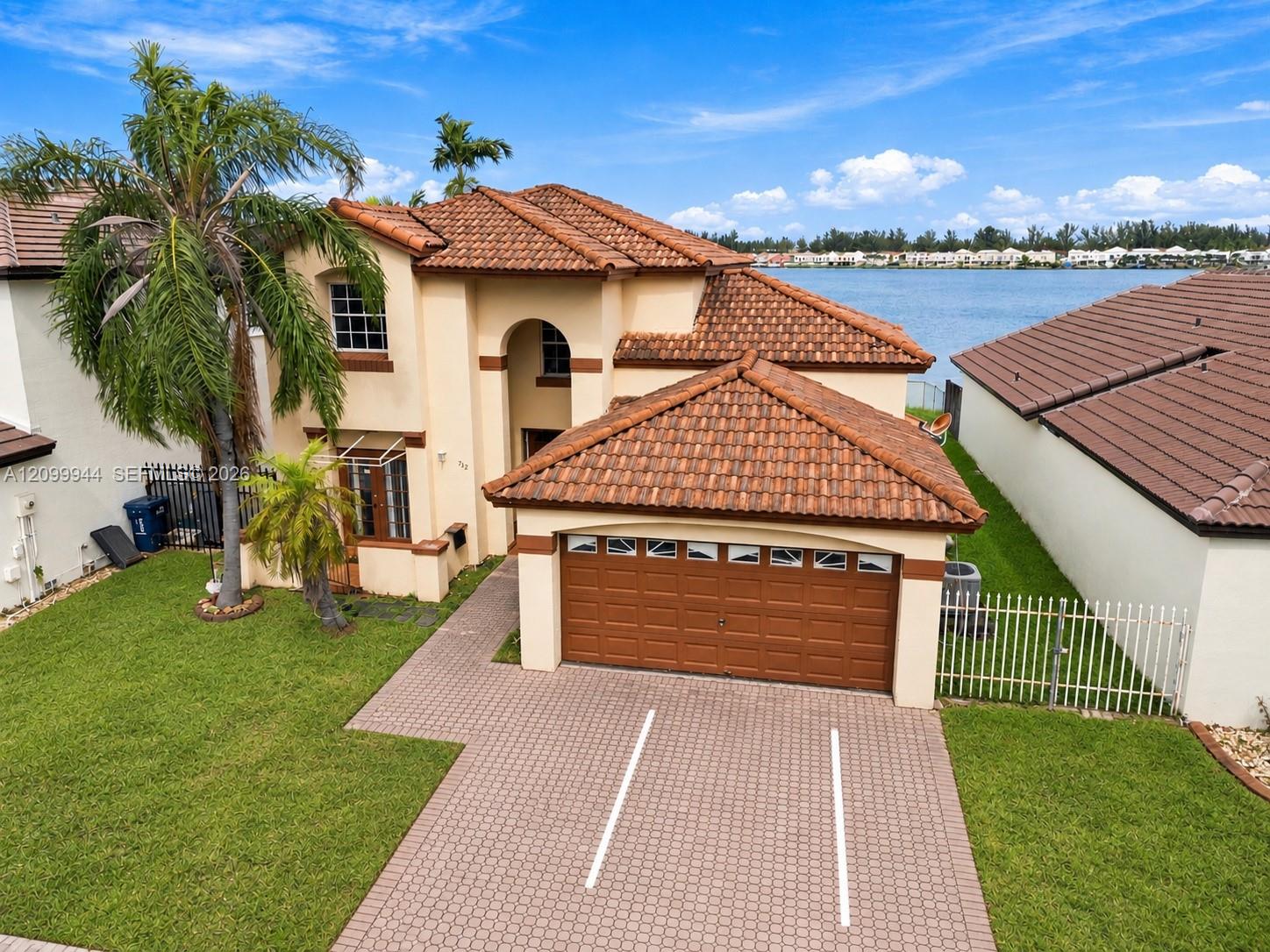 Waterfront two-story stucco home with terracotta tile roof, palm trees, and a paver driveway.