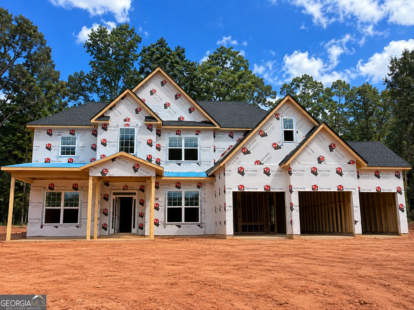 Two-story home under construction with steep gables, exposed housewrap, and an unfinished dirt lot.