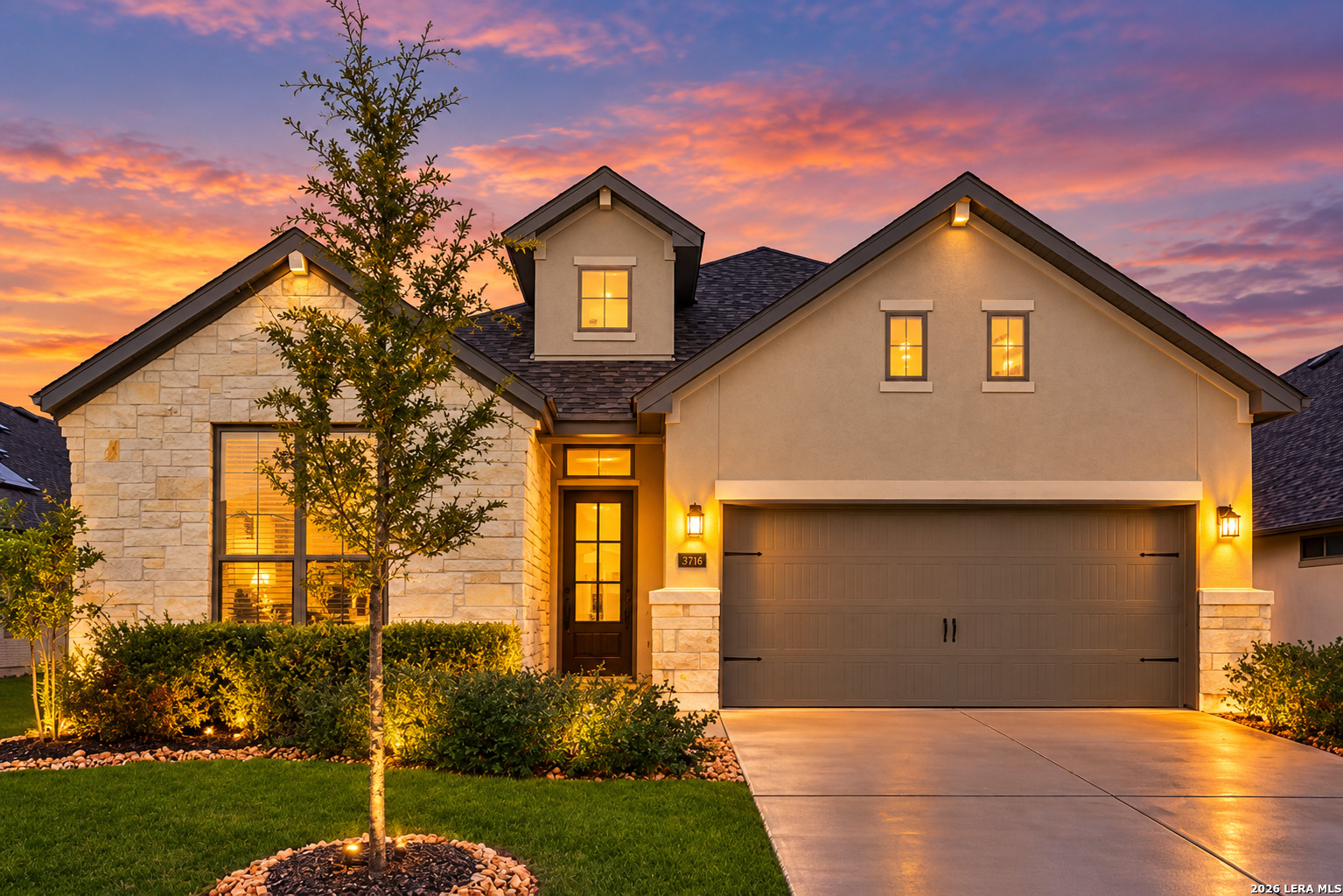 Two-story stone-and-stucco home with illuminated windows, landscaped lawn, and broad driveway at dusk.