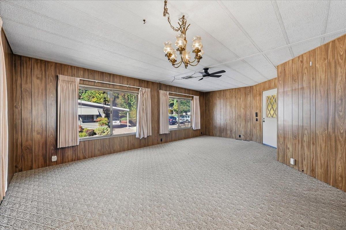 Empty carpeted room with wood-paneled walls, two curtained windows, and a chandelier.