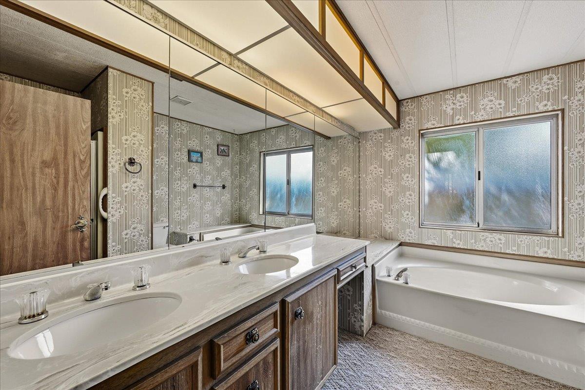 Bathroom with a long double-sink vanity, mirrored wall, patterned wallpaper, and white bathtub.