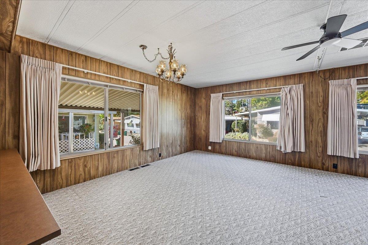 Empty bedroom with wood-paneled walls, light carpet, curtained windows, chandelier, and ceiling fan.