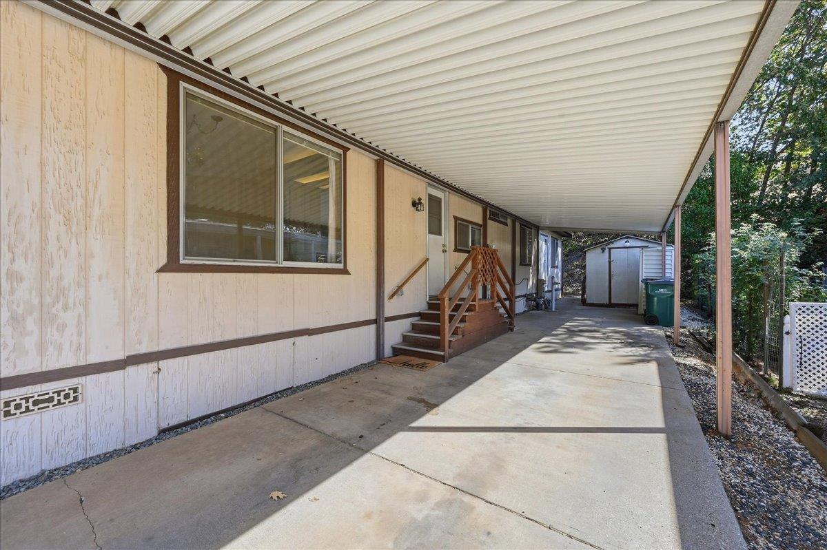 Covered concrete carport beside a light-colored home, with wooden entry steps and a shed at the far end.