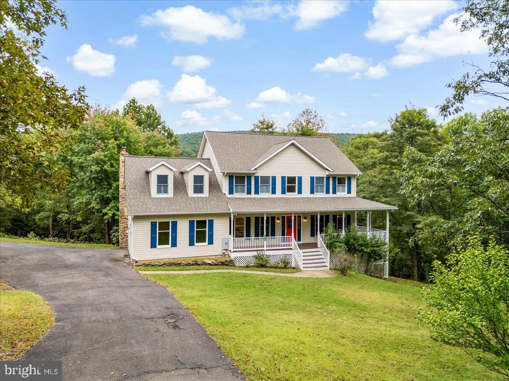 Two-story beige house with blue shutters, a covered porch, and a long driveway amid mature trees.