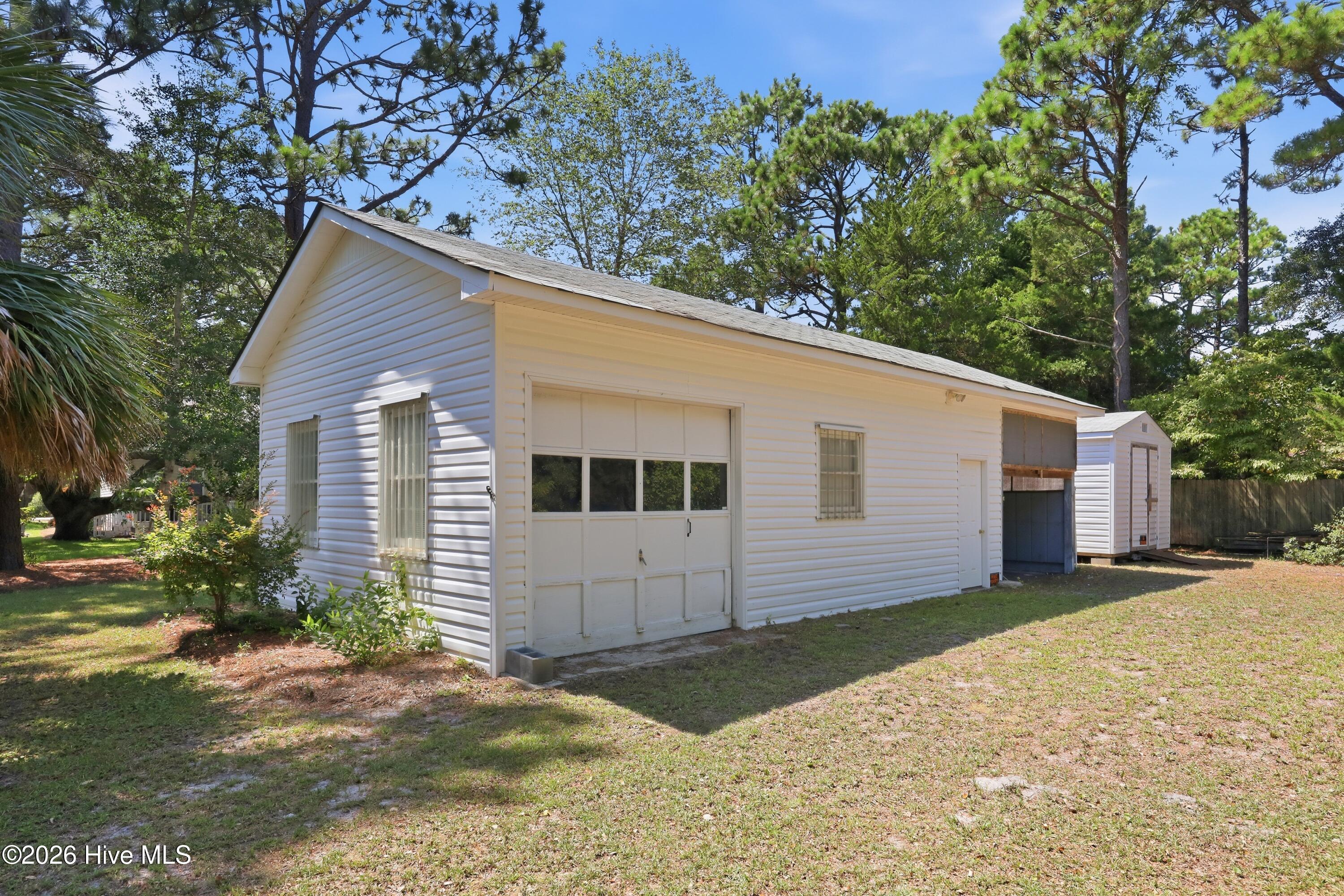 White detached garage with a large paneled door and a small shed behind it.