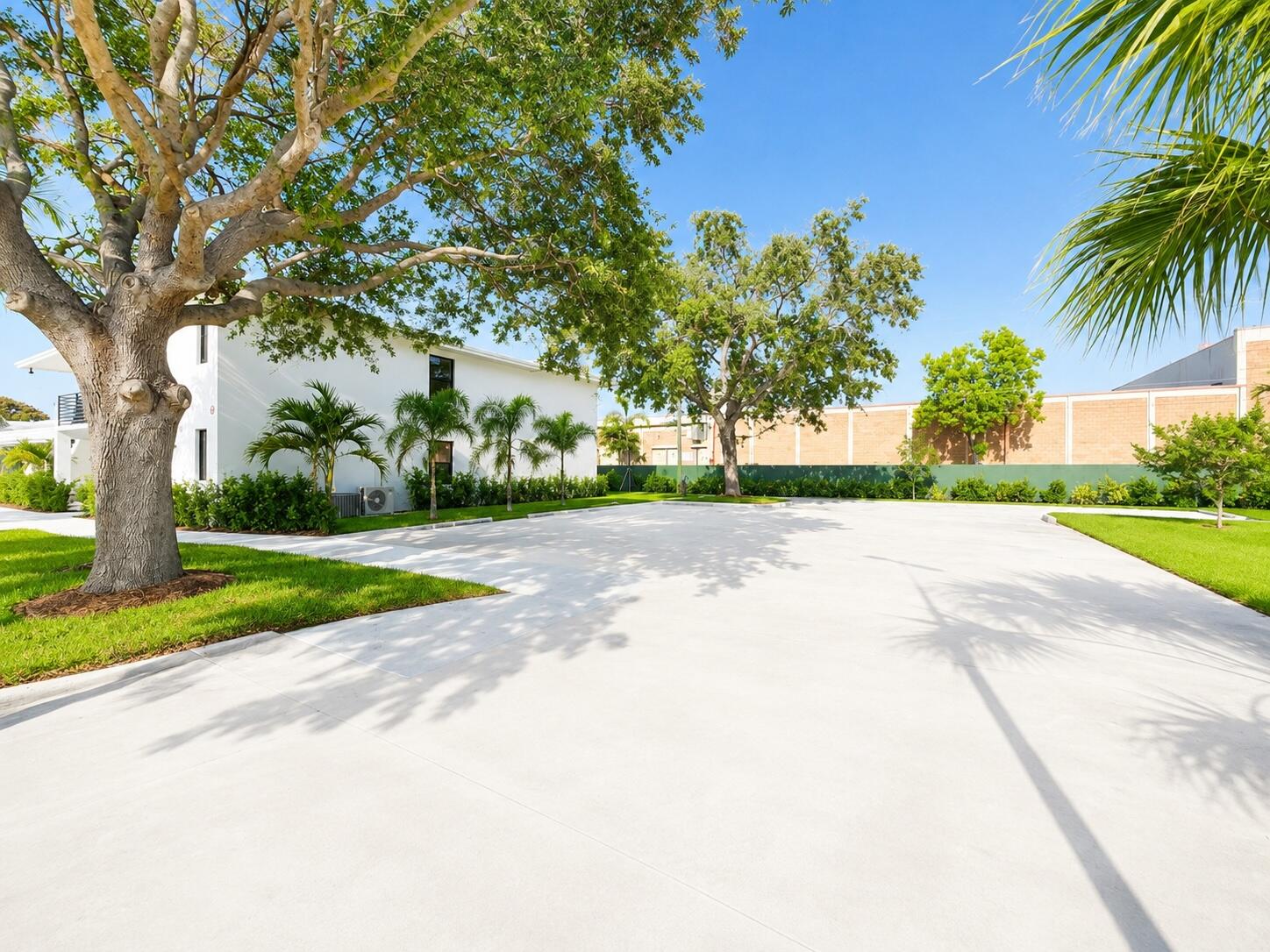White building beside a broad paved parking area shaded by mature trees and palms.