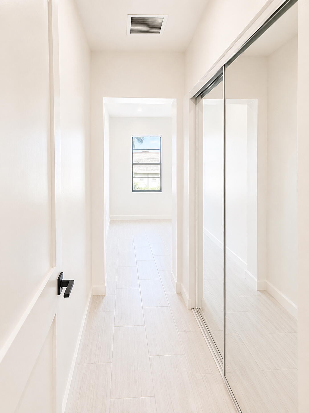 Bright hallway with pale tile flooring, mirrored sliding closet doors, and a window at the far end.
