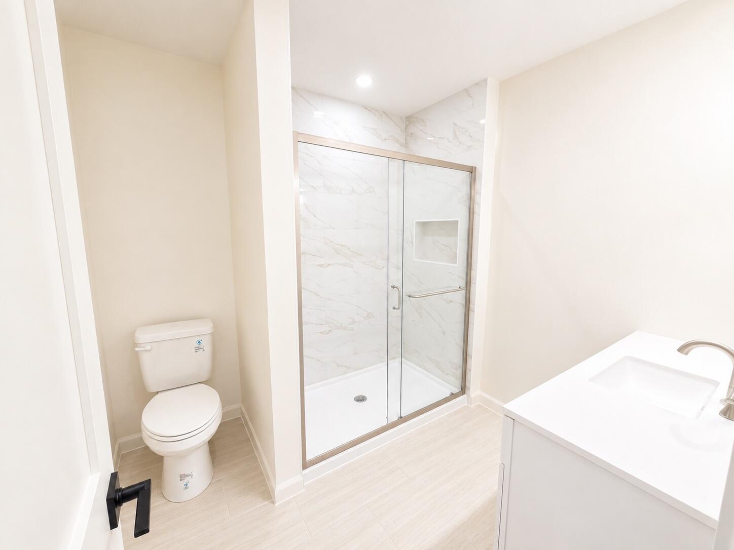 Bathroom with a glass shower lined in marble-look tile beside a toilet and white vanity.