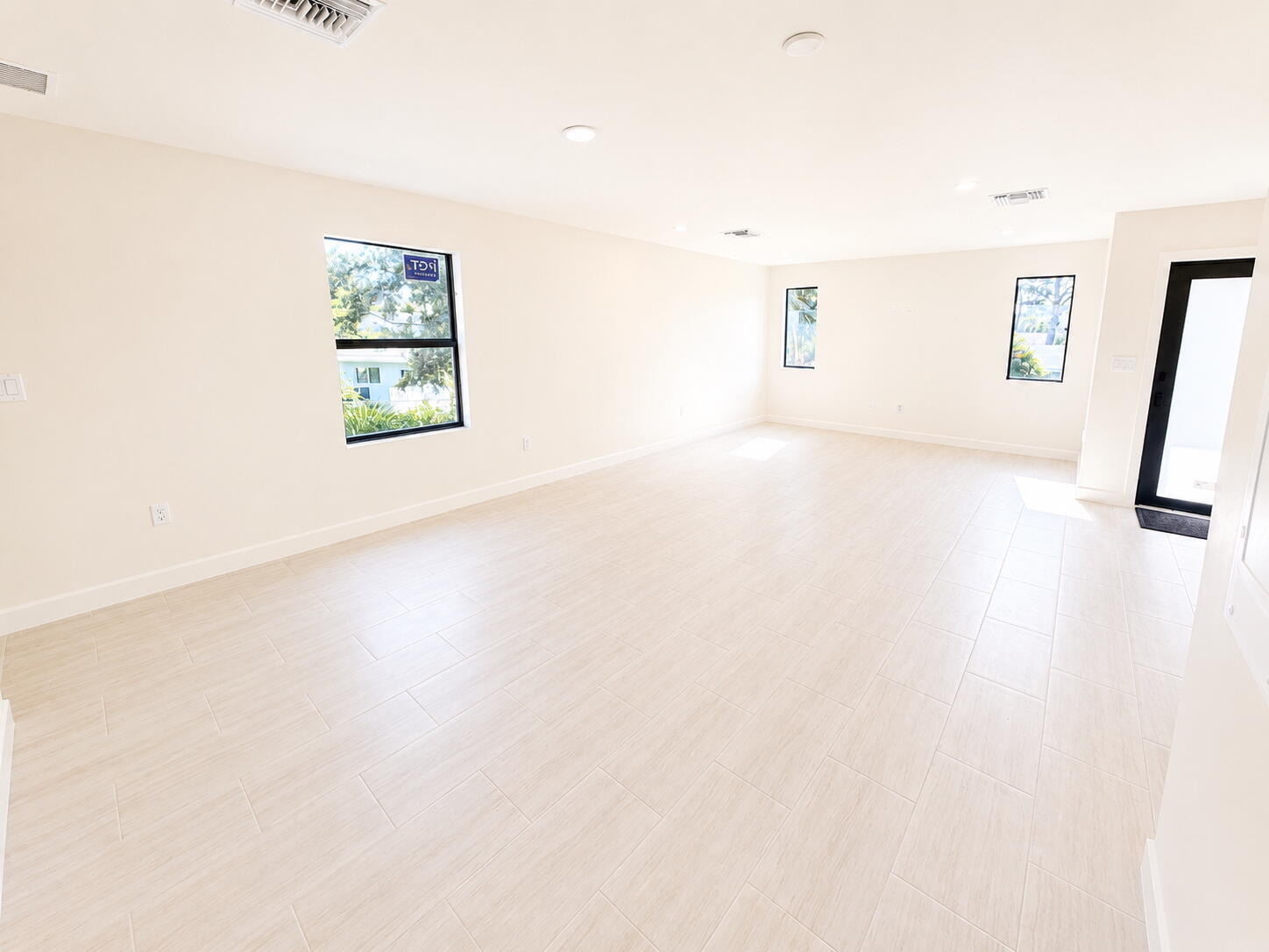 Empty living area with pale tile floors, three windows, and a black-framed glass entry door.