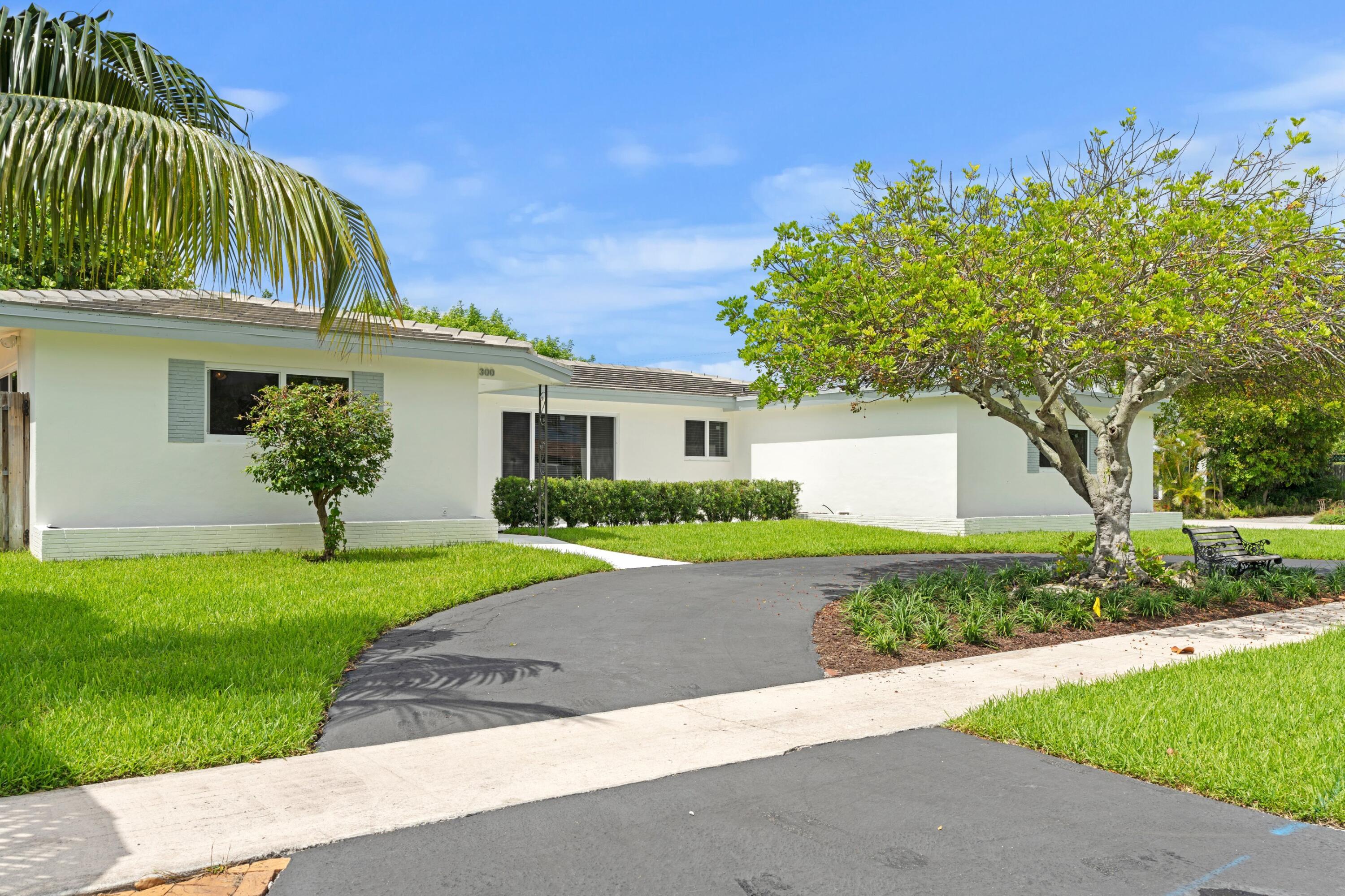 White single-story home with a curved driveway, manicured lawn, and mature tropical trees.