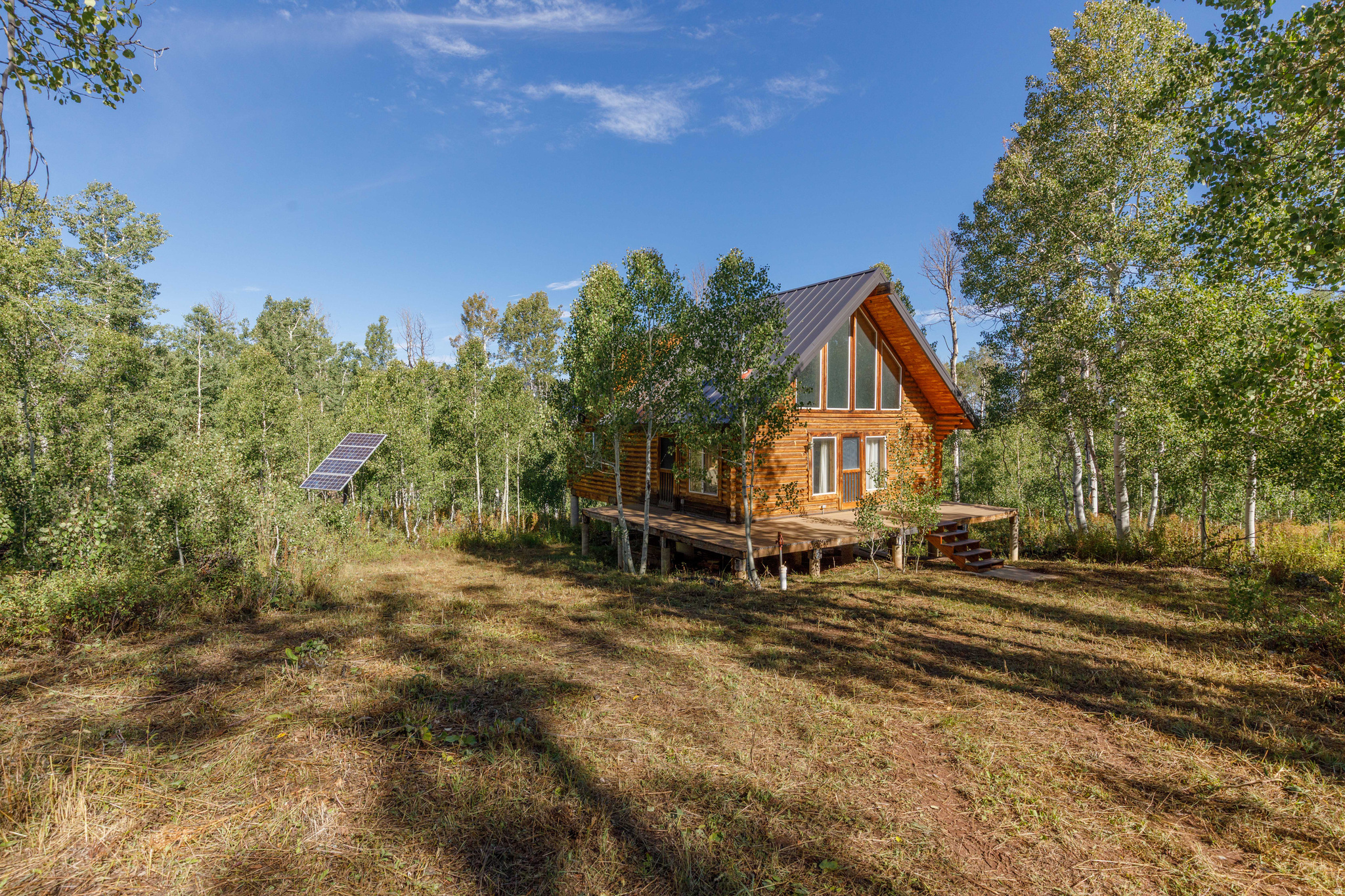 Log cabin in a wooded clearing, with a steep roof, raised deck, and nearby solar panel.