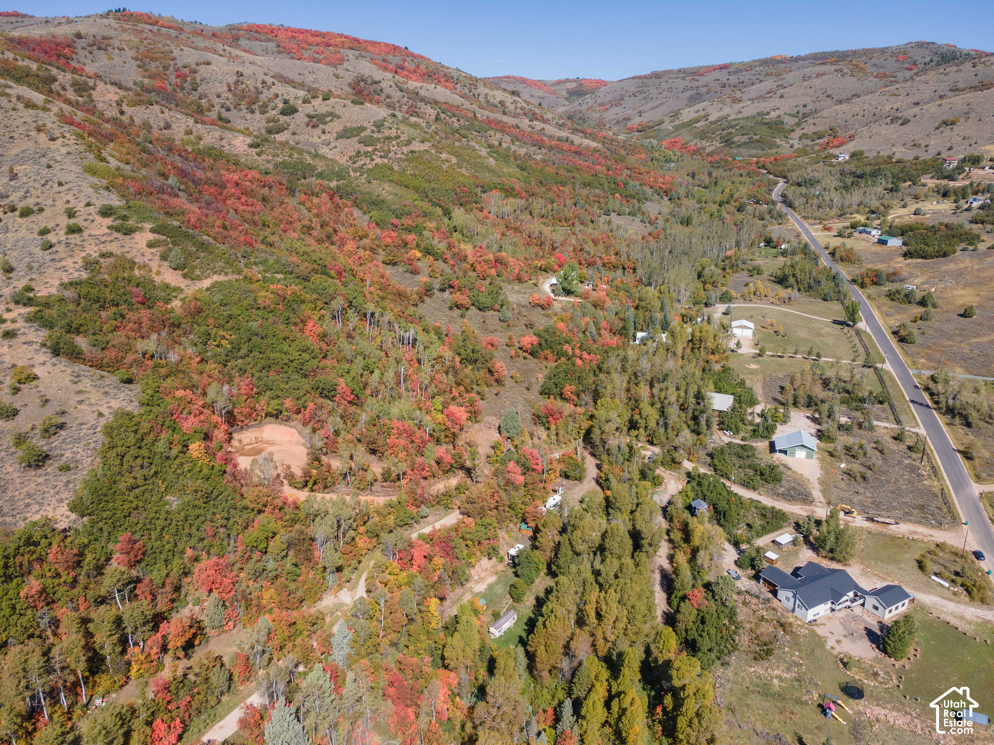 Aerial view of a rural home and wooded valley in autumn, with colorful hillsides and a winding road.