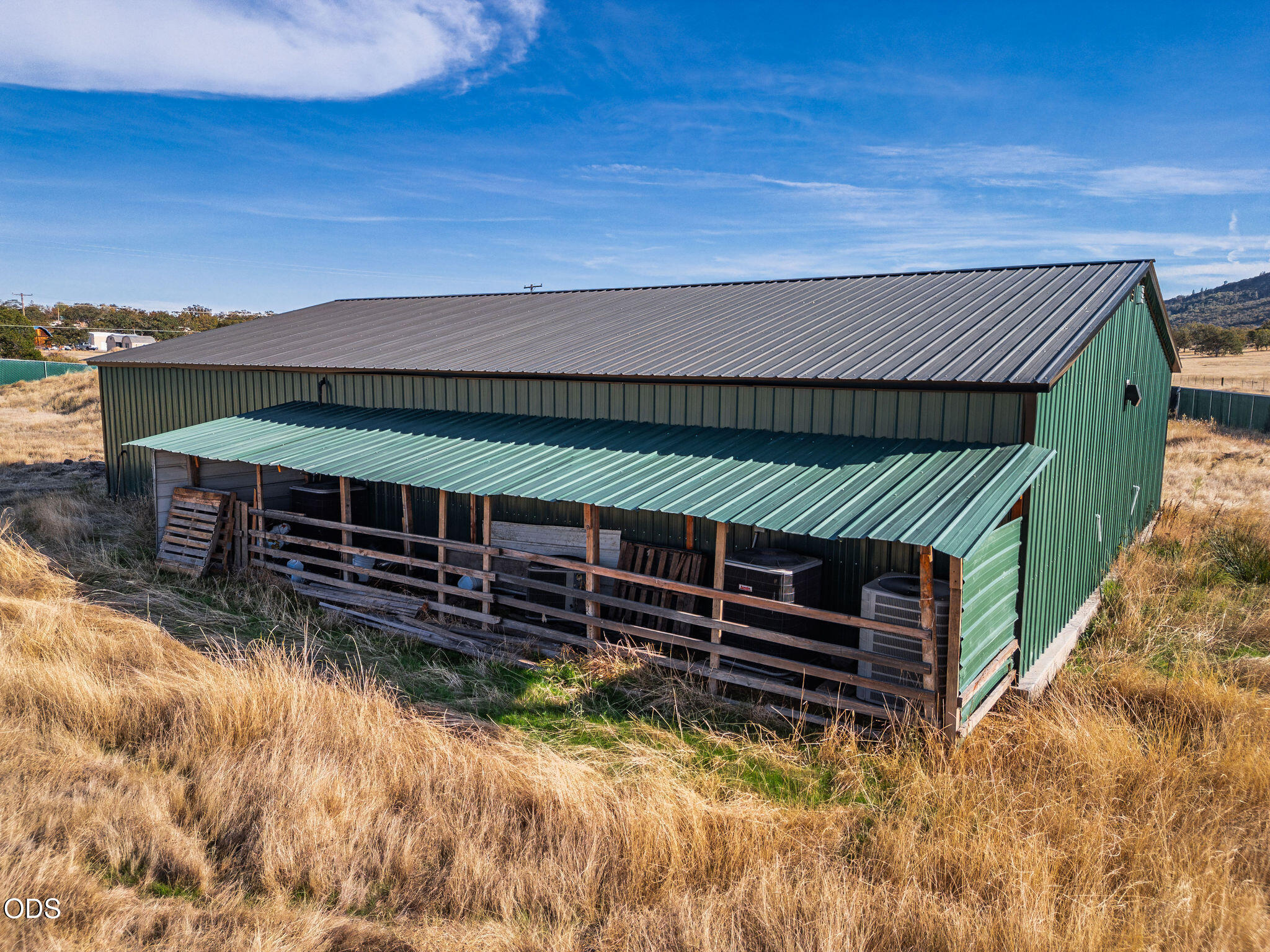 Green metal barn with a covered, fenced enclosure in dry grass.