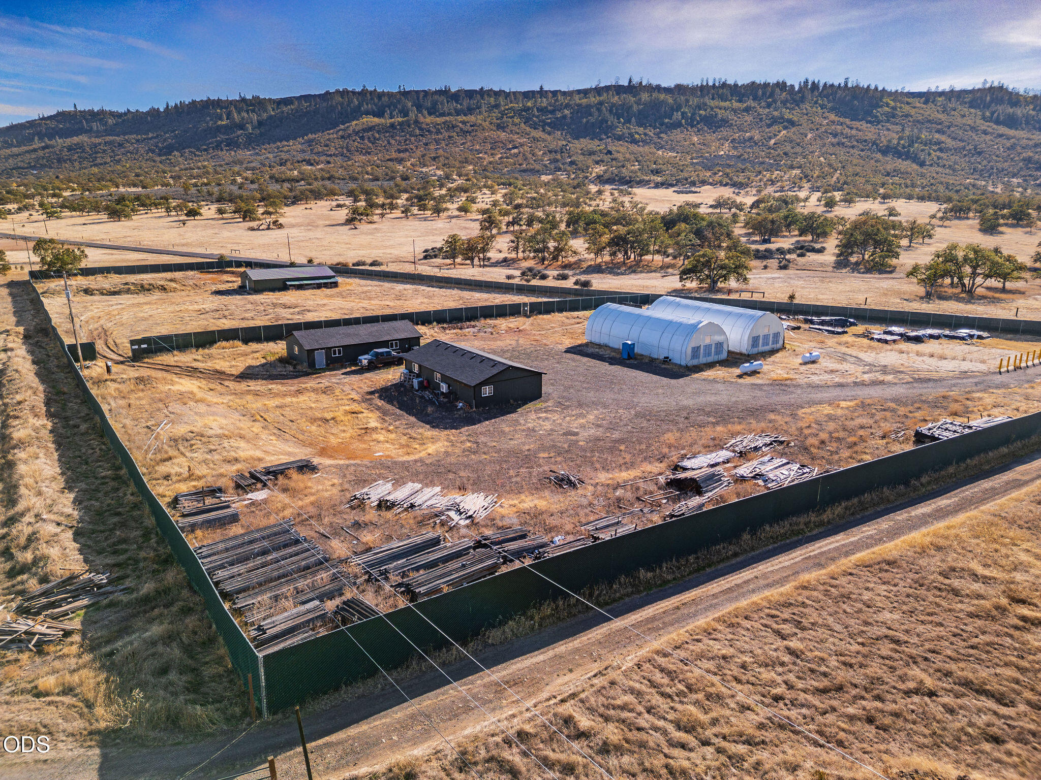 Fenced rural property with hoop greenhouses, two small buildings, and wooded hills beyond.