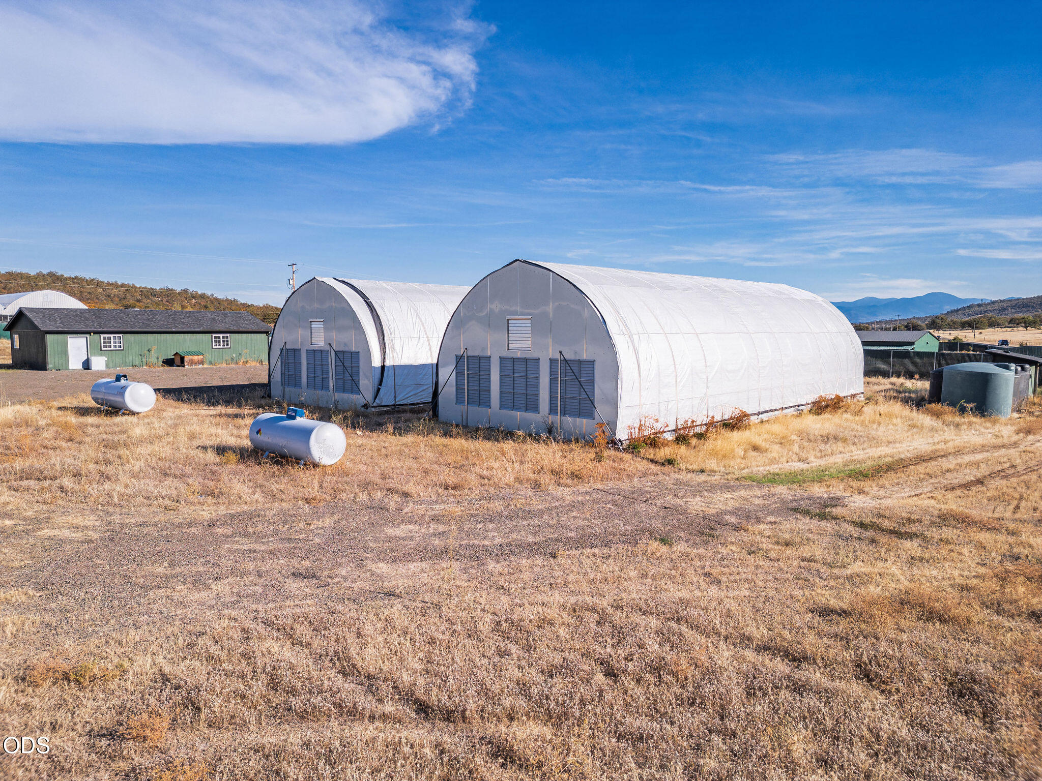 Two large white arched agricultural buildings stand in a dry field with mountains in the distance.