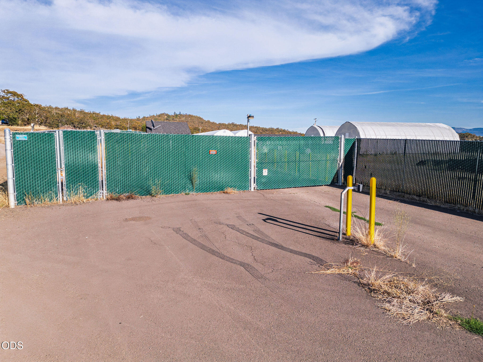Paved property entrance with a green-screened fence, black gate, yellow bollards, and hoop-house greenhouses.