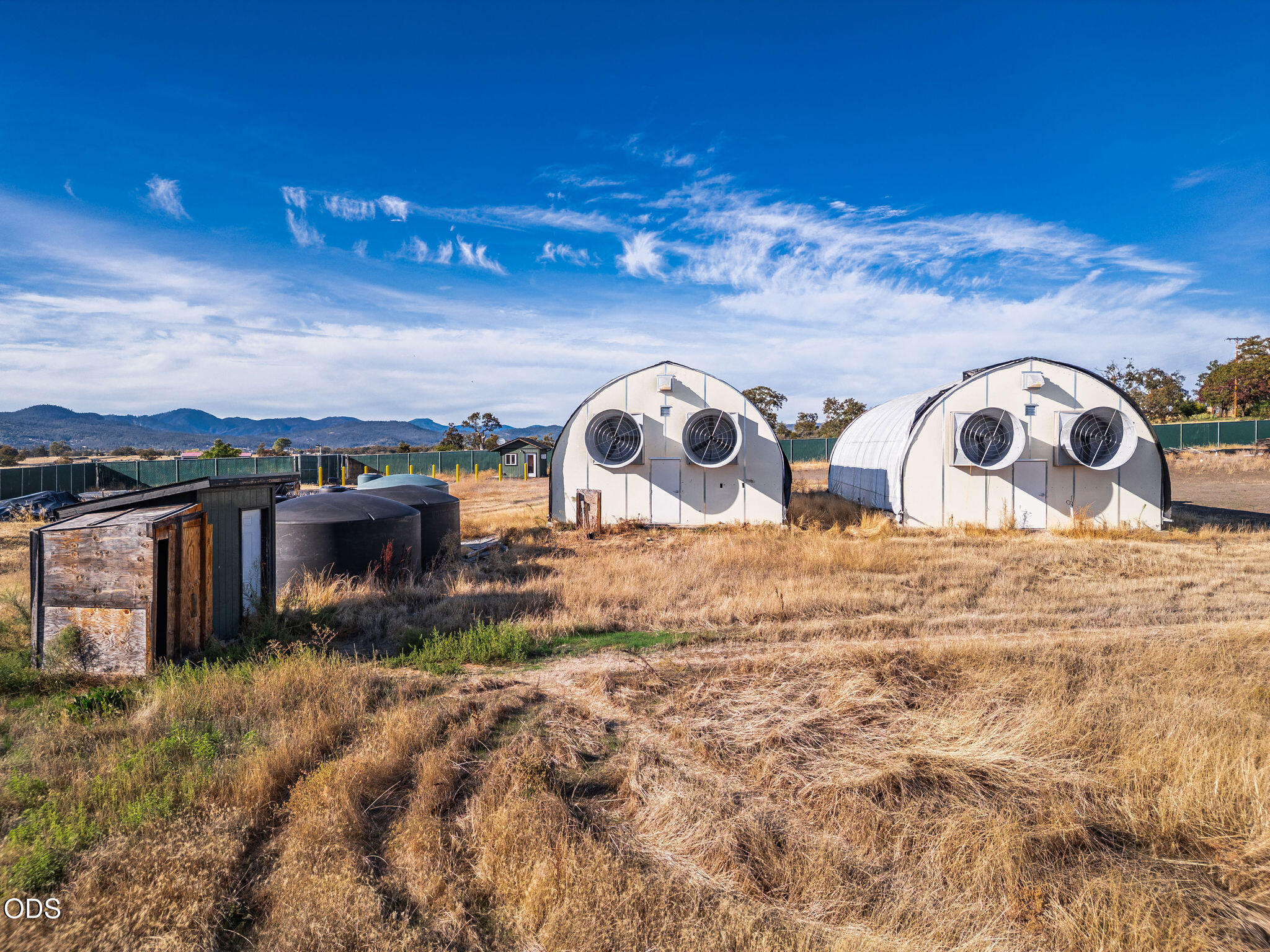 Two arched agricultural buildings with large ventilation fans on a dry, grassy property.