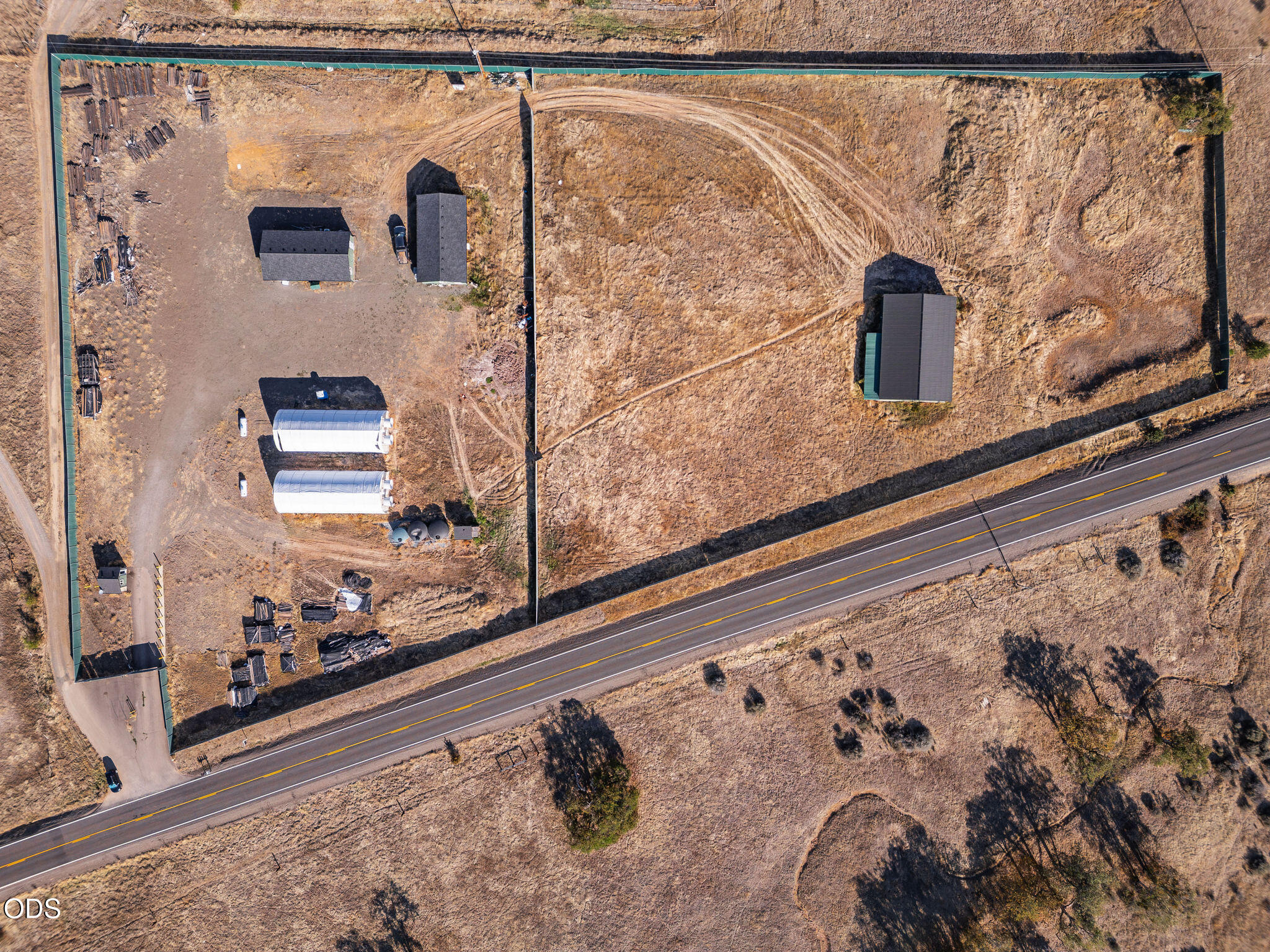 Aerial view of fenced rural property with scattered outbuildings beside a paved road