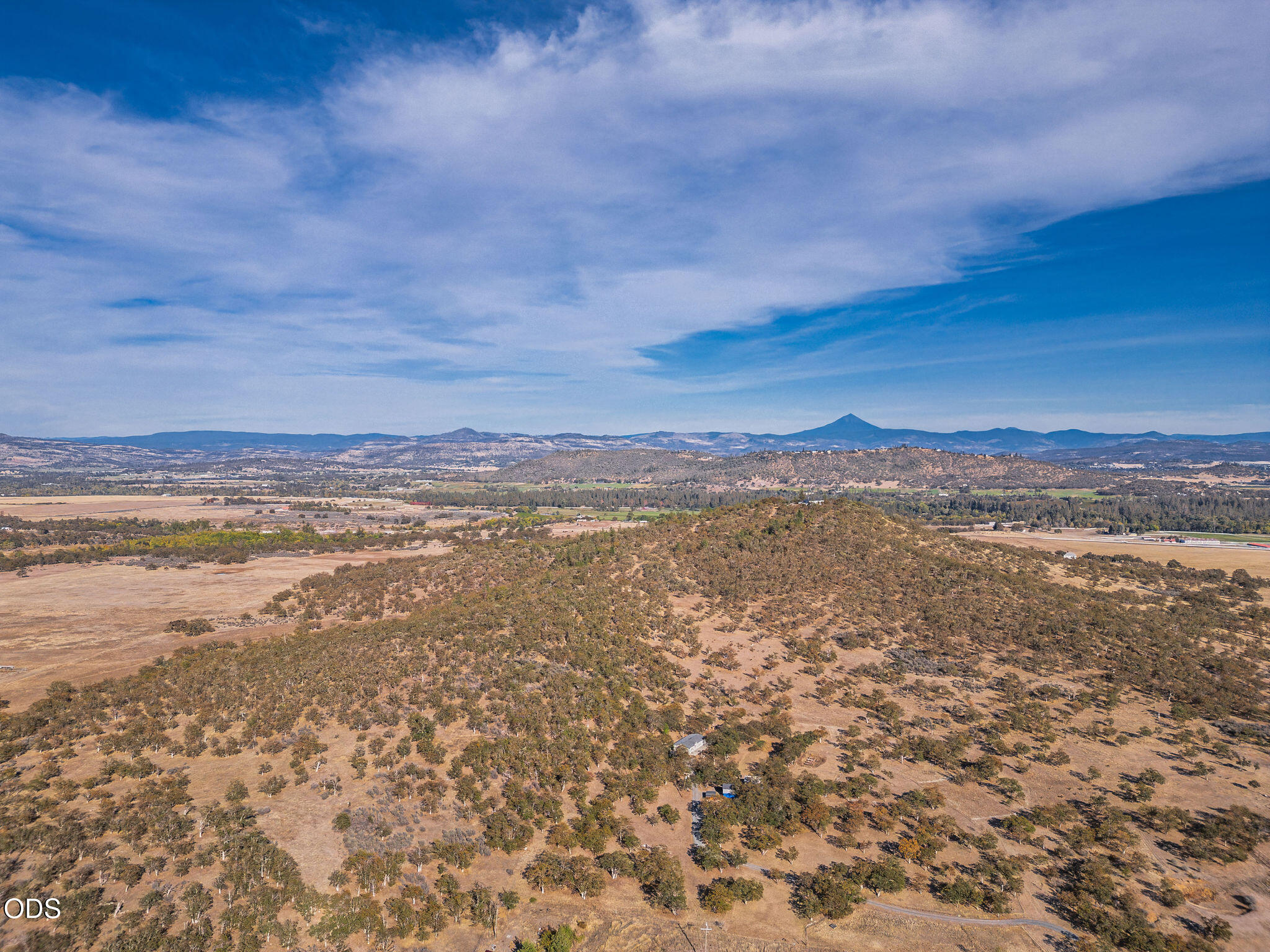 Aerial view of dry, tree-dotted acreage with small structures and distant mountains.