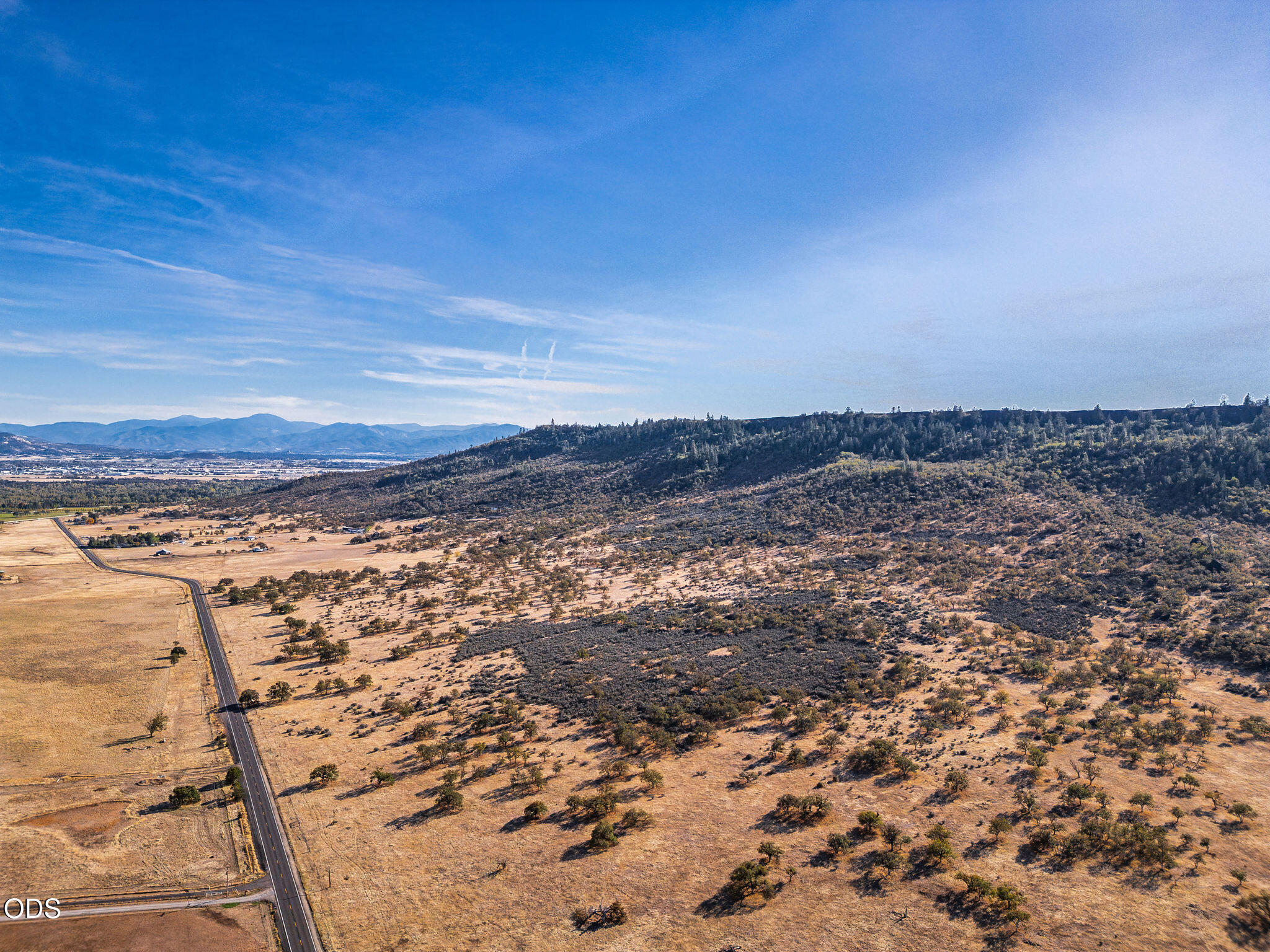 Aerial view of dry acreage with a road, scattered trees, wooded hills, and distant mountains.
