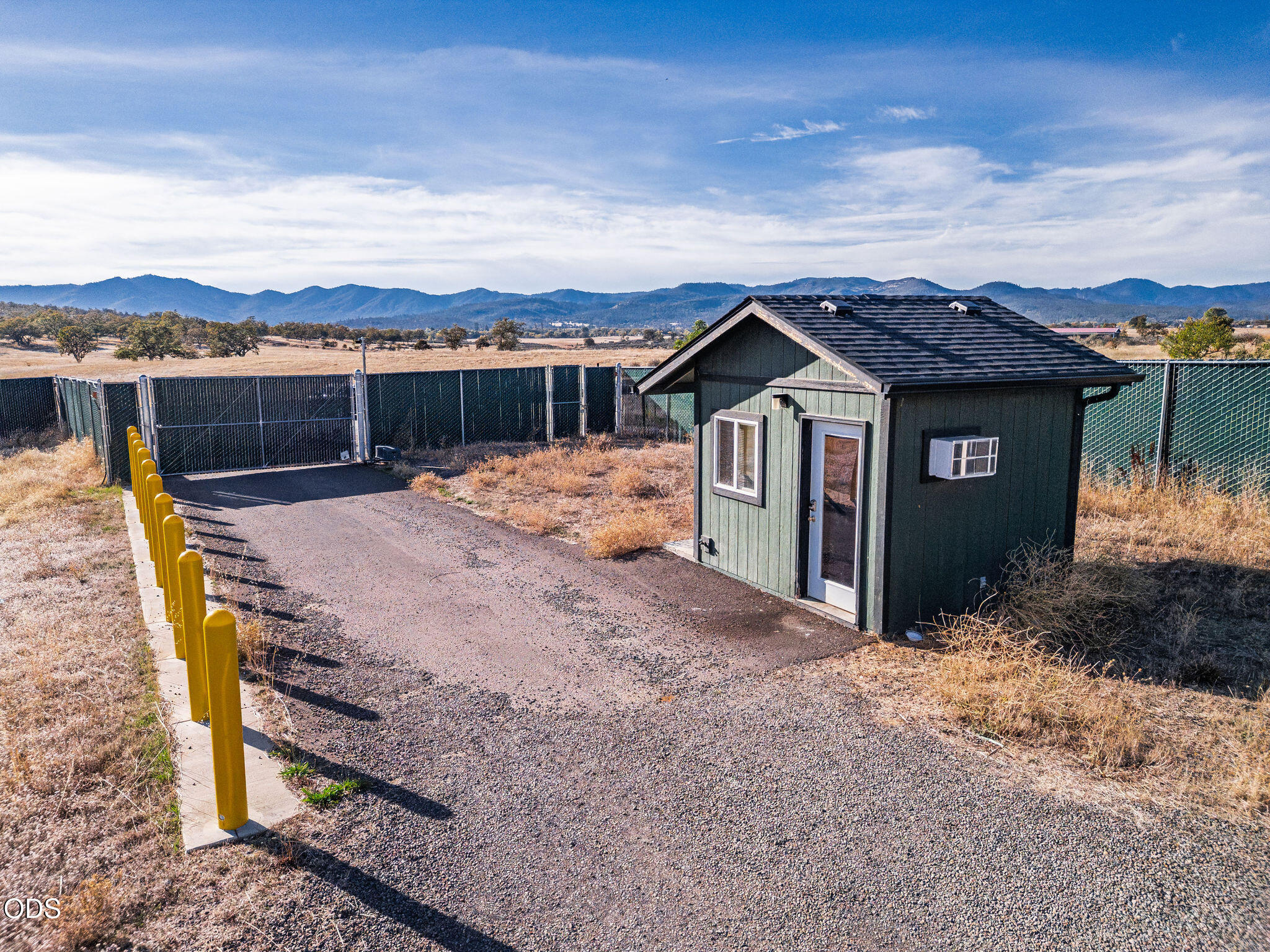 Small green outbuilding beside a fenced gravel area, with mountain views.