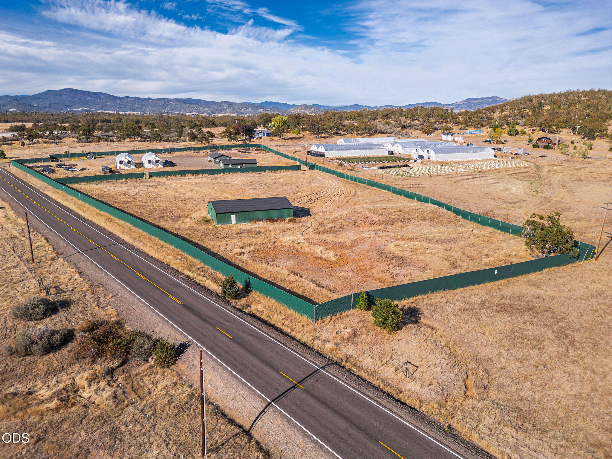 Fenced rural property beside a road, with greenhouse tunnels and mountain views.
