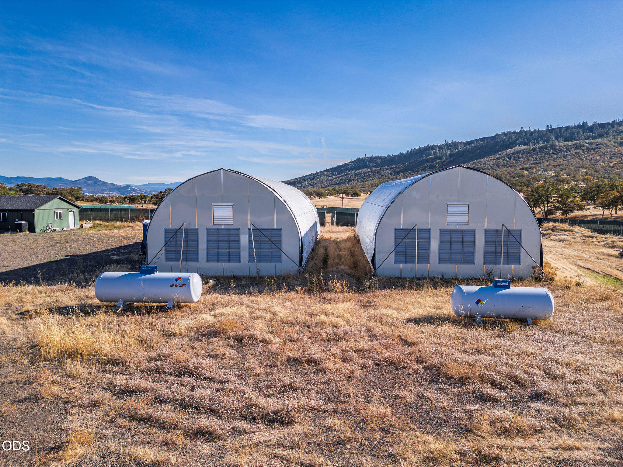 Two white arched agricultural buildings and propane tanks in a dry field with wooded hills beyond.