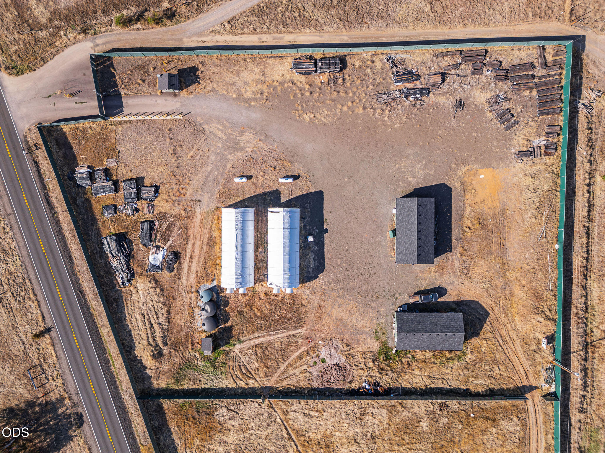 Aerial view of fenced storage yard with lumber stacks, covered structures, and roadside access.
