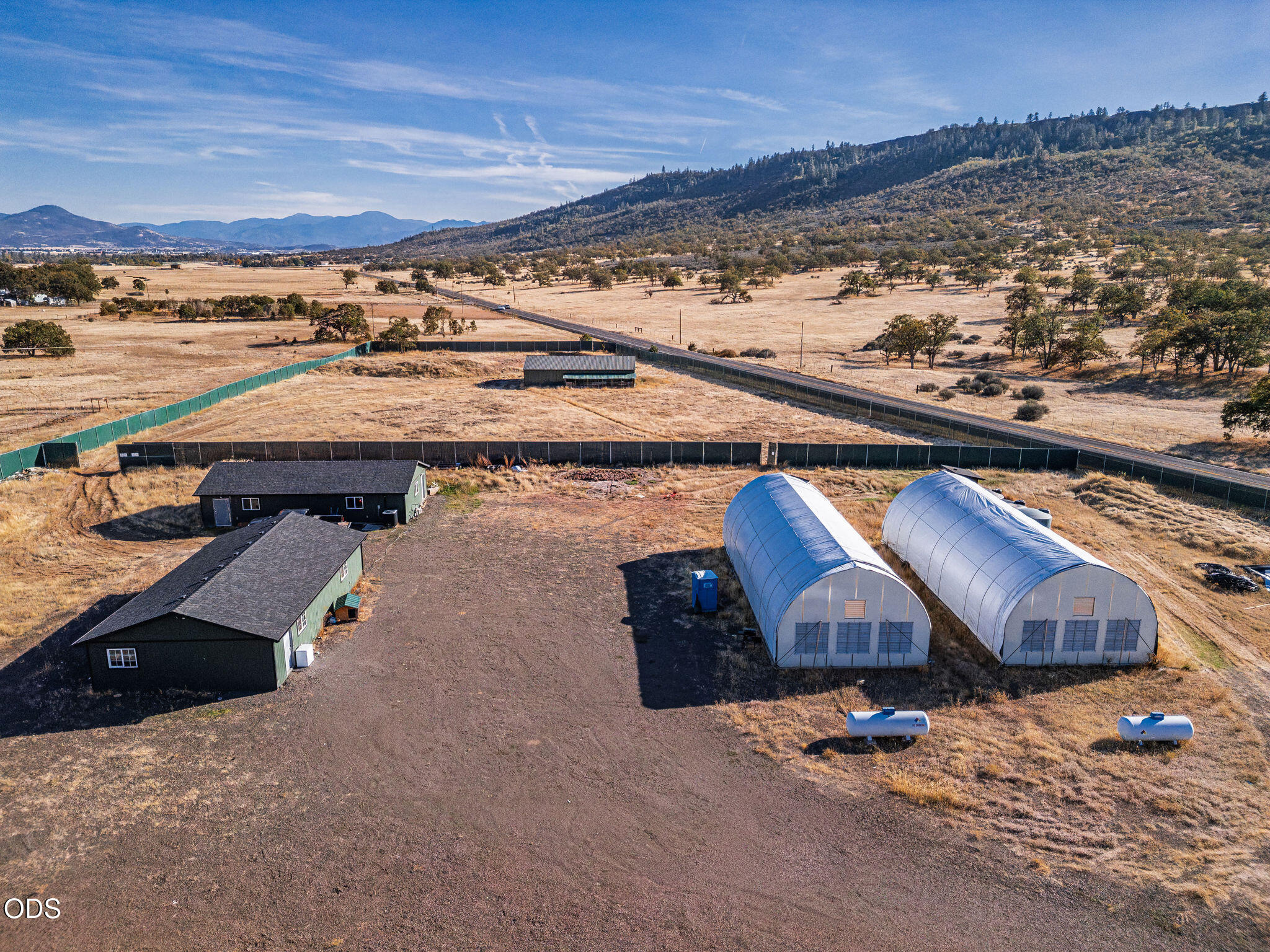 Aerial view of a rural property with two buildings and two hoop greenhouses beside dry fields and wooded hills.