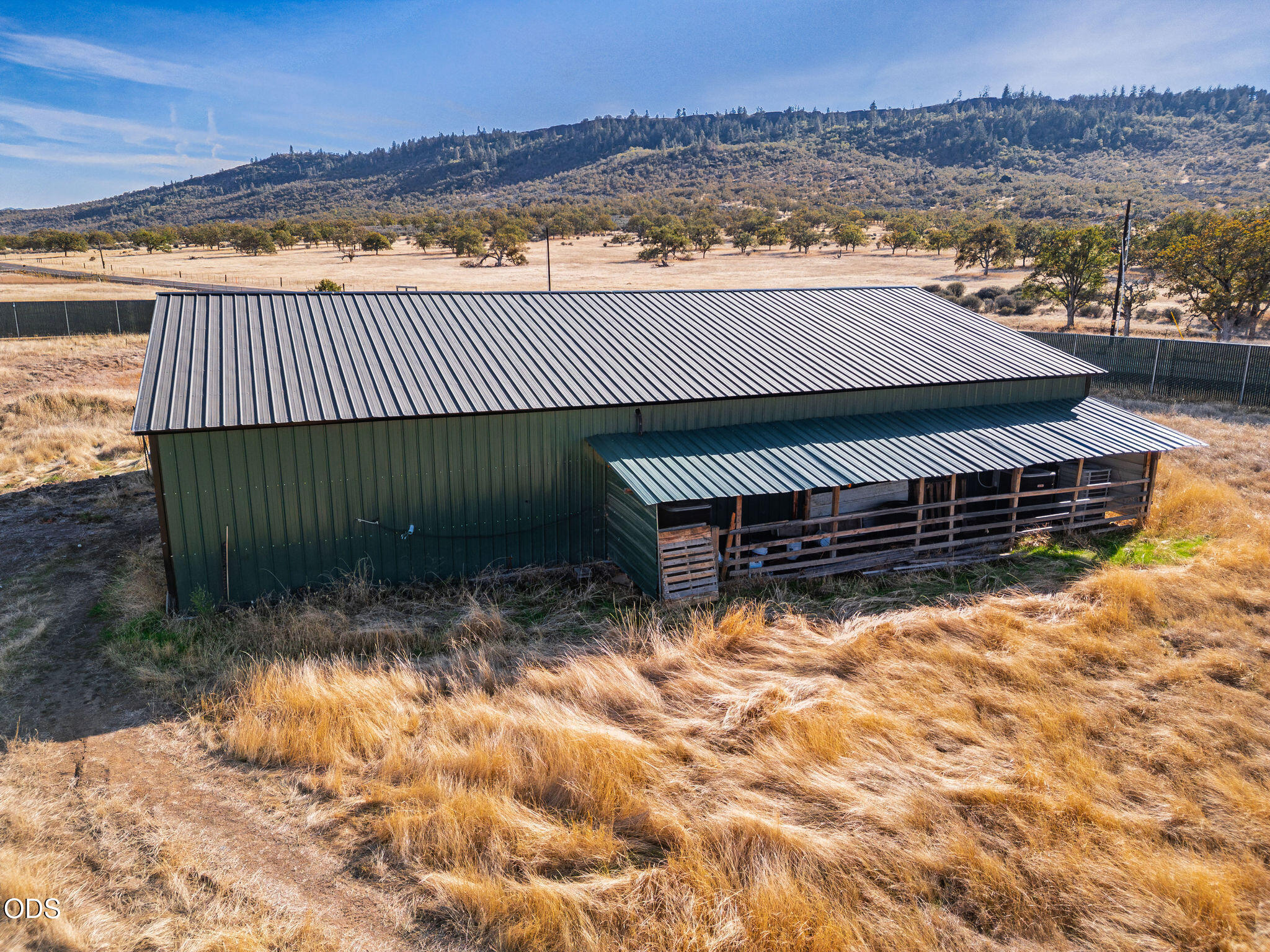 Dark green metal barn with a covered livestock pen, surrounded by dry grassland and wooded hills.