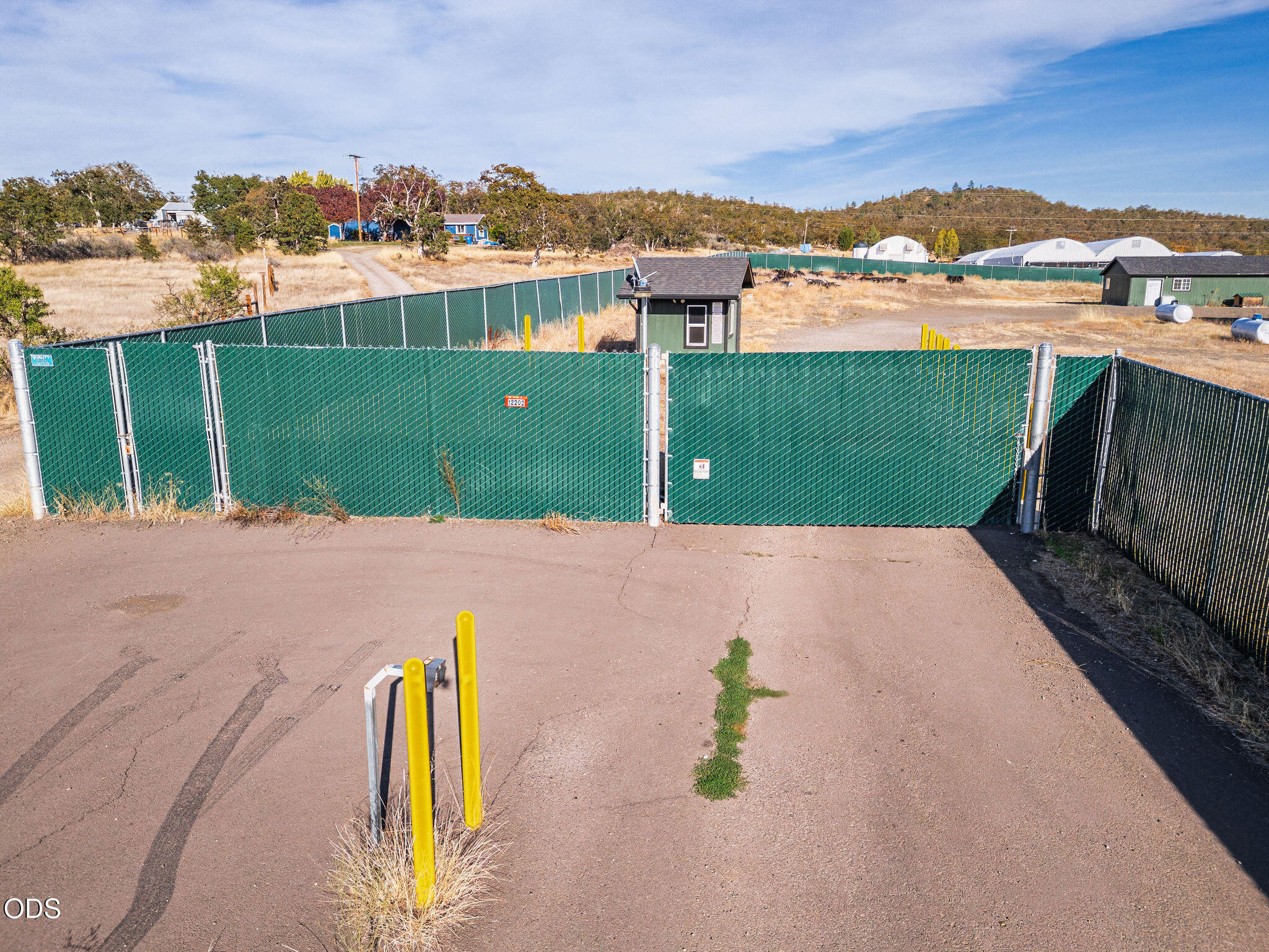 Fenced property entrance with green chain-link gate, paved driveway, yellow bollards, and a small guard booth.