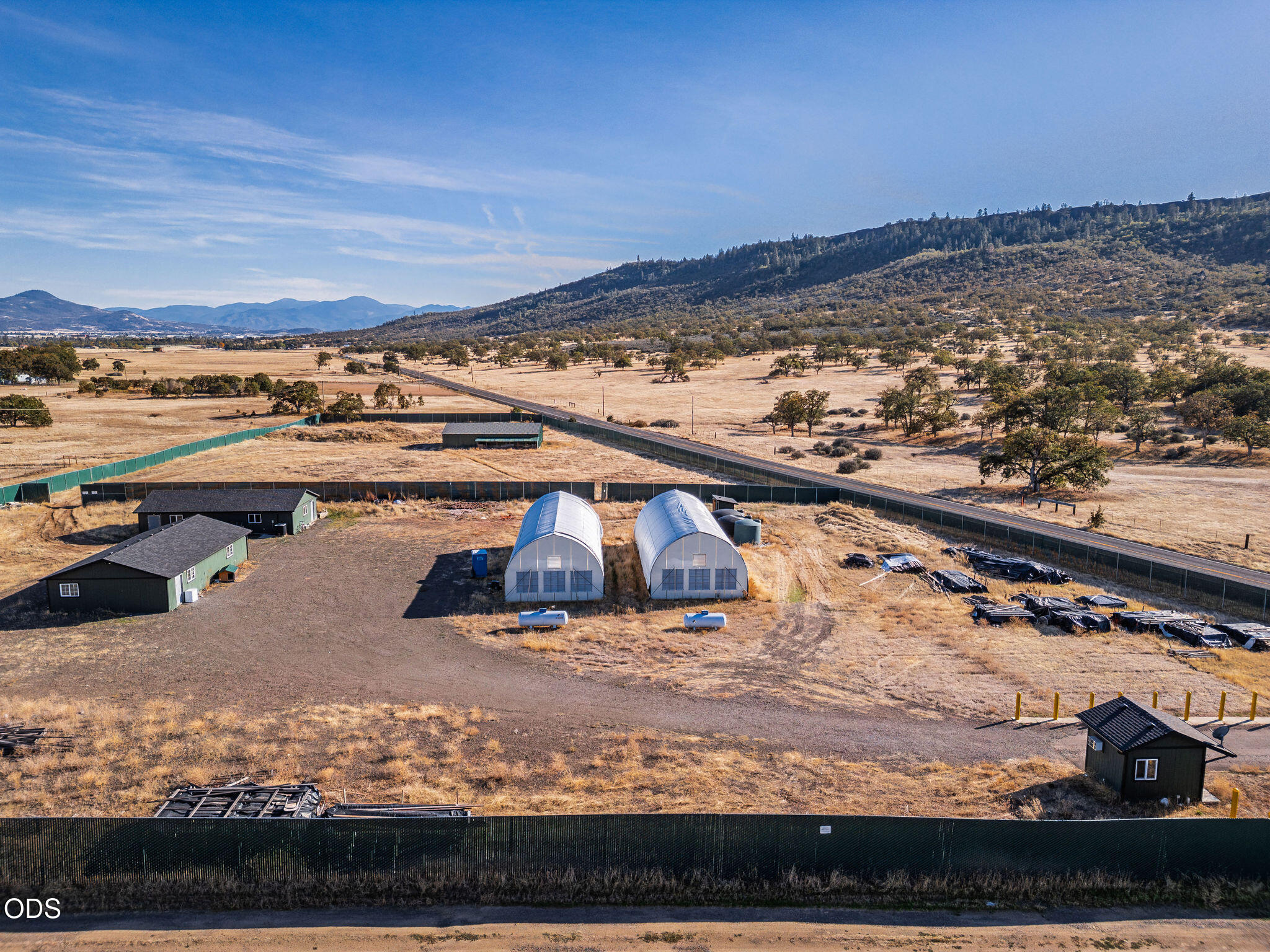 Aerial view of a fenced rural property with two hoop-house greenhouses and a wooded mountain ridge beyond.