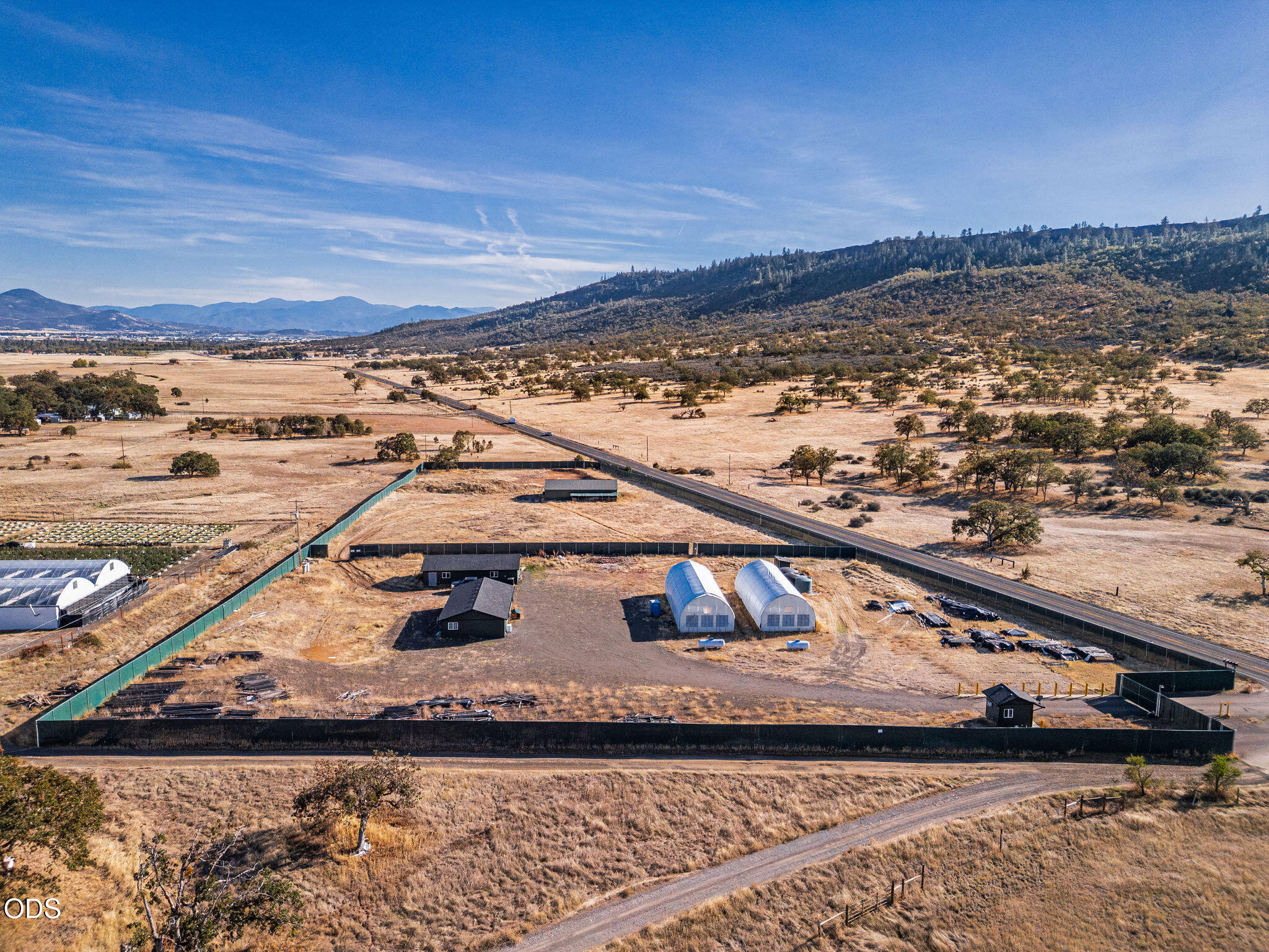 Aerial view of a rural property with two greenhouses, small buildings, and mountains beyond.