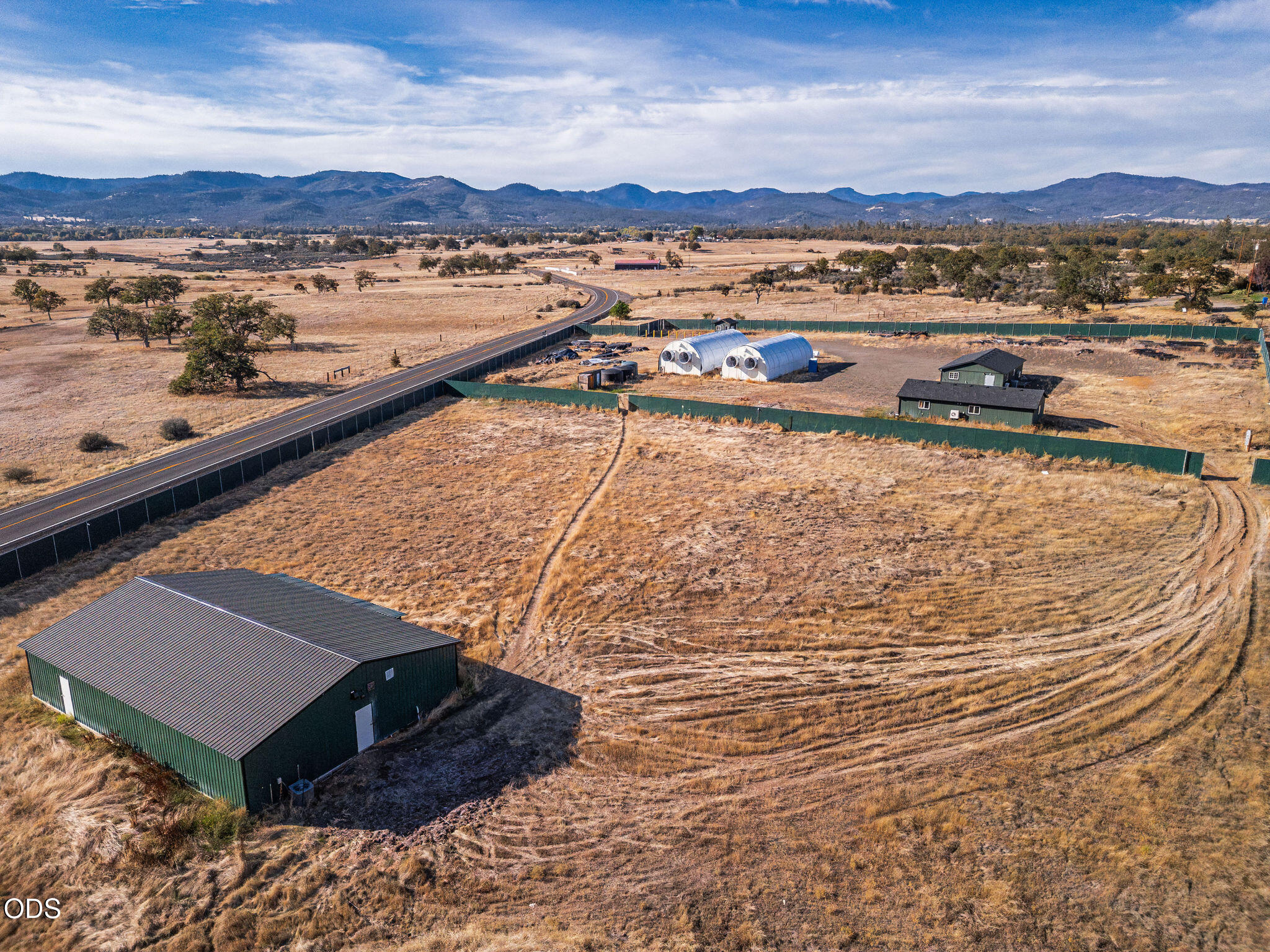 Aerial view of fenced rural acreage with a green house, metal outbuilding, and two white arched structures.