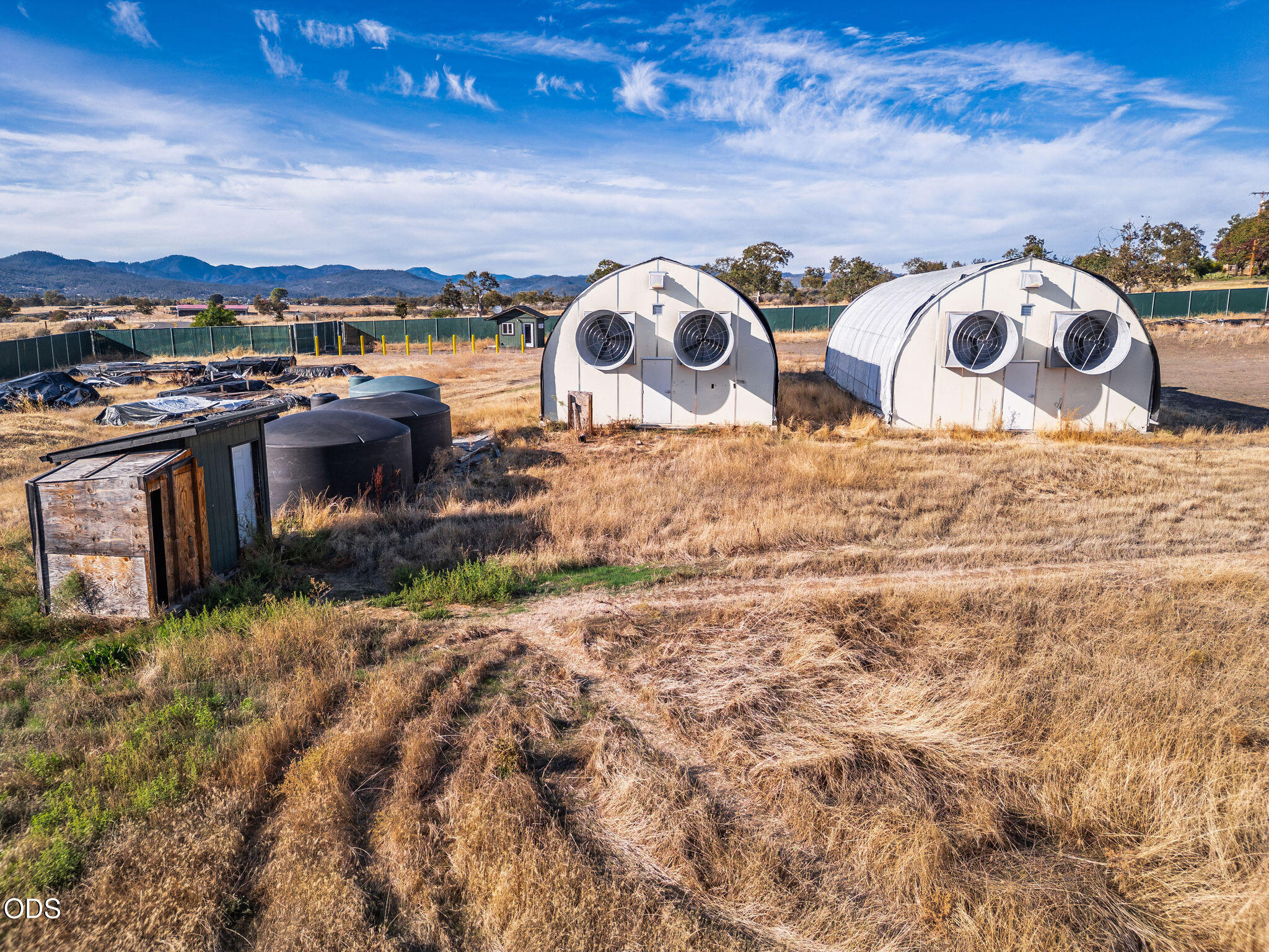 Rural property with two fan-ventilated arched buildings, water tanks, and dry grass against distant mountains.