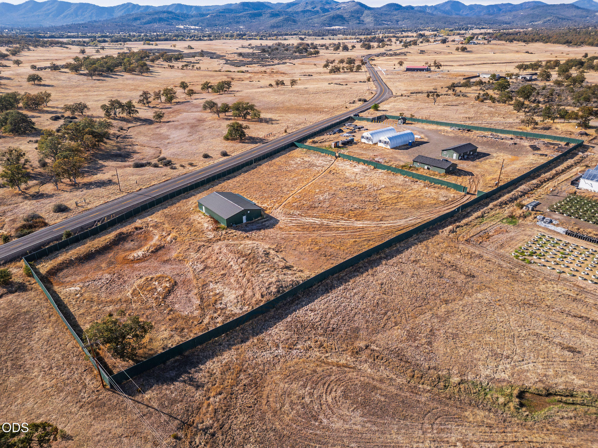 Fenced rural property with outbuildings and hoop houses, surrounded by dry fields and distant mountains.